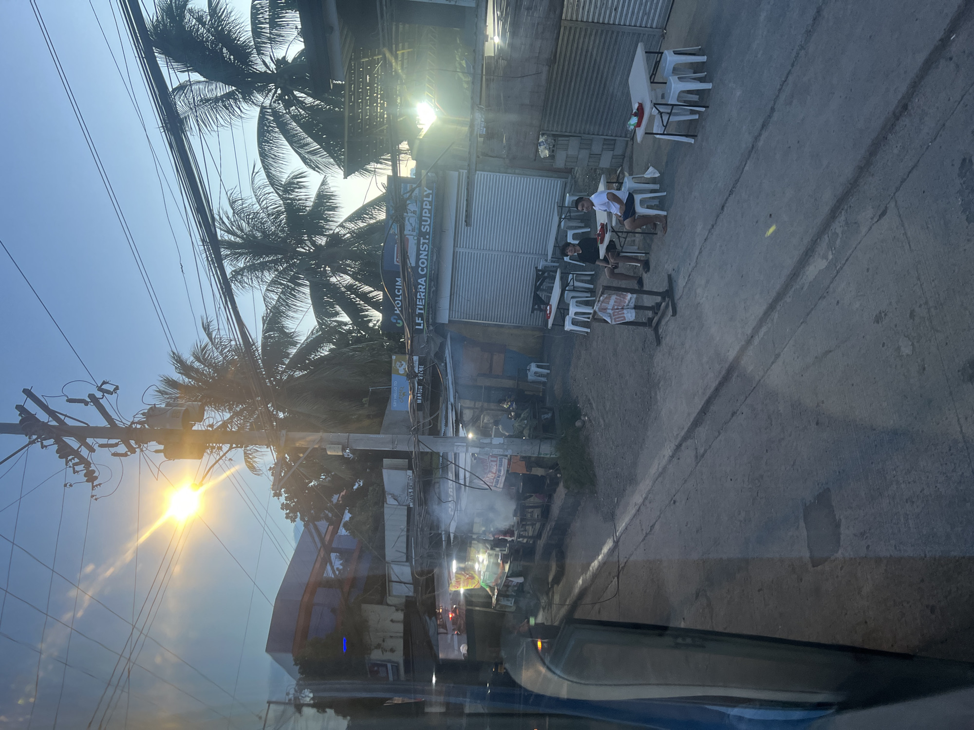 The image depicts a street scene in Davao City, Philippines, characterized by a mix of commercial establishments and residential structures. The time of day appears to be late afternoon or early evening, with the sun casting long shadows and the sky displaying hues of blue and orange. Several individuals are visible, some seated at tables outside a small eatery, while others are likely engaged in work or daily activities within the shops. The presence of numerous utility poles and electrical wires suggests an urban or semi-urban setting. The architecture is a blend of simple, tin-roofed structures and more substantial concrete buildings, indicative of ongoing development and diverse economic activities. The overall atmosphere suggests a busy, informal commercial area with a sense of community.
