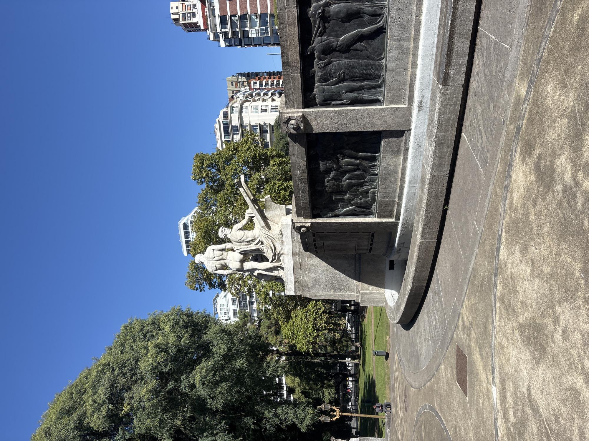 This image captures a grand stone monument situated in a public park in Buenos Aires, Argentina, under a clear, bright blue sky. The monument features prominent light-colored statues at its top, depicting two classical figures. One figure stands tall, appearing to hold a long, vertical object, while the other stands nearby, looking toward the left.
The lower sections of the monument are adorned with dark, detailed bas-relief panels. These panels depict numerous figures in various poses, suggesting historical or allegorical scenes. The monument itself is constructed from large blocks of stone or concrete, showing texture and some weathering.
Surrounding the monument is a paved plaza area, which transitions into a verdant park. Lush green trees dominate the midground and foreground, providing ample shade. In the distance, through the trees, several modern multi-story residential buildings are visible, adding to the urban backdrop.
The scene is bathed in strong, natural sunlight, indicating it is likely midday or early afternoon. Clear skies prevail, contributing to the bright and open atmosphere. In the park area, near a decorative lamppost, two individuals can be seen sitting on a bench. There is no visible text legible on the monument or in the immediate surroundings.