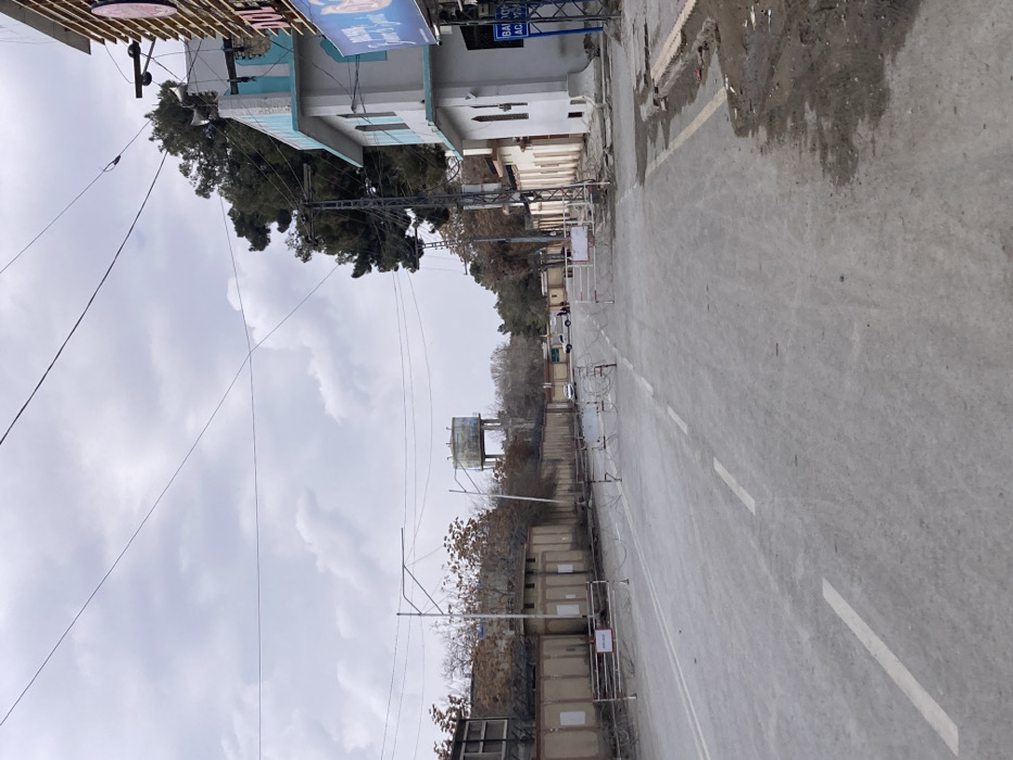 The image captures a street scene in Quetta City Tehsil, Pakistan, under an overcast sky. The asphalt road, marked with white dashed and solid lines, extends into the distance. In the midground, multiple metal barriers and coils of concertina wire are deployed across the road, effectively blocking vehicular passage. Several barriers display red and white "No Entry" signage.
On the left side of the road, a long, low-rise building with a beige-colored exterior and multiple roof-mounted fixtures is visible, bordered by trees, some with exposed branches suggesting a winter or dry season. Further down the road on the left, an elevated, weathered water tank on a concrete stand is prominent against the grey sky.
On the right side of the road, a two-story building features light grey walls with distinctive turquoise-blue accents on its upper facade. A commercial sign, partially visible, indicates "DUL" at the top and "BALOCH ACADEMY" lower down. Adjacent to this building on the far right, a structure with a vertically striped brown facade is present. The unpaved shoulder on the immediate right appears dusty and uneven.
Numerous utility poles and a complex network of overhead power lines crisscross the frame. In the far distance, beyond the roadblocks, a white pickup truck or van is parked on the road, and a few individuals are standing on the right-hand side of the street near the vehicle.