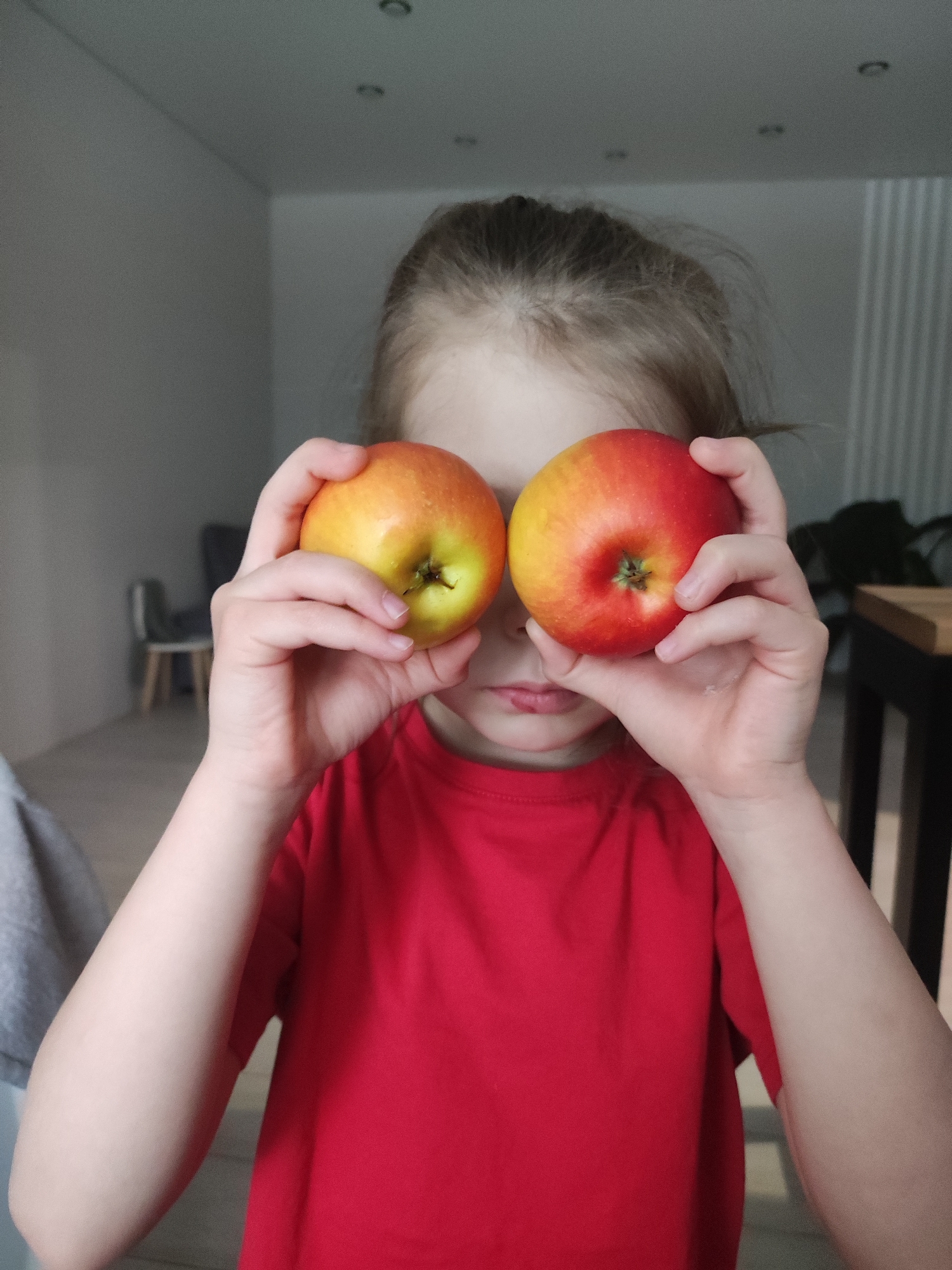 A young child holds two apples in front of their eyes. The child has fair skin, light brown hair pulled back, and wears a red t-shirt. One apple is predominantly yellow with some red, while the other is predominantly red. The background shows a sparsely decorated room with light-colored walls, a light-colored floor and minimal furniture. The location is unknown and cannot be confirmed to be Votkinsk, Russia.