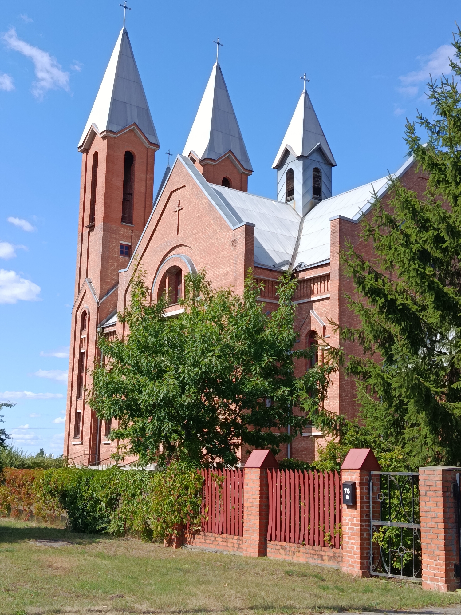 The image shows a red brick church with a tall bell tower and two smaller spires on a clear, sunny day. The church has a metal roof and a prominent cross atop each spire. A red wooden fence with brick pillars encloses the property, and a metal gate is visible on the right side. There are green trees and bushes surrounding the church, and the ground is covered with grass. The scene appears to be in a rural or suburban area. The address number "76" is visible on a mailbox attached to one of the brick pillars. This church is likely located in the city of Svietlahorsk, Belarus.