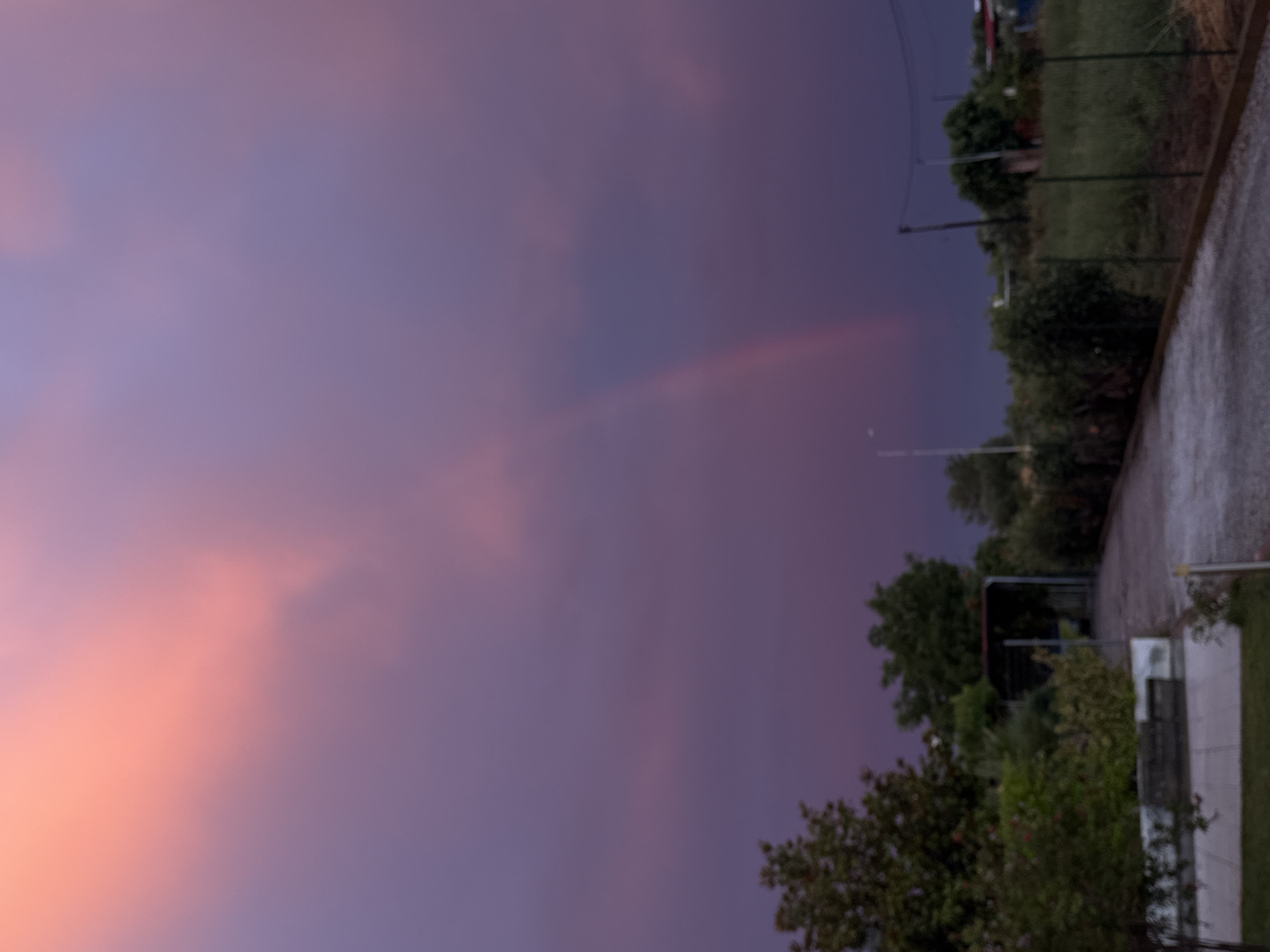 The image depicts a landscape scene at dusk, as indicated by the soft, gradient colors of the sky transitioning from warm pinks and oranges near the horizon to deep purples and blues overhead. A faint rainbow is visible arching across the purple part of the sky. The foreground features a paved area, possibly a driveway or path, with some greenery and trees on the right side. Beyond the greenery, there appears to be a fence and more vegetation, suggesting a semi-rural or suburban setting. A few light poles or thin structures are also present among the trees. The lighting suggests it is either sunrise or sunset. There are no people visible in the image. The overall mood is serene and atmospheric, characteristic of twilight.
