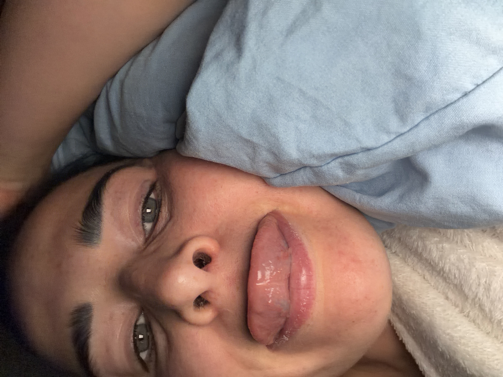 A close-up, rotated selfie-style photograph shows a person lying down with a blue blanket partially covering their head. The person's face is the primary focus, displaying grey eyes, dark eyebrows, a nose with prominent nostrils, and lips slightly parted with the tongue visible. The background includes a white, possibly textured surface beneath the person. There is no indication of the location being Saint Petersburg, Russia or any specific city. The scene appears to be indoors.