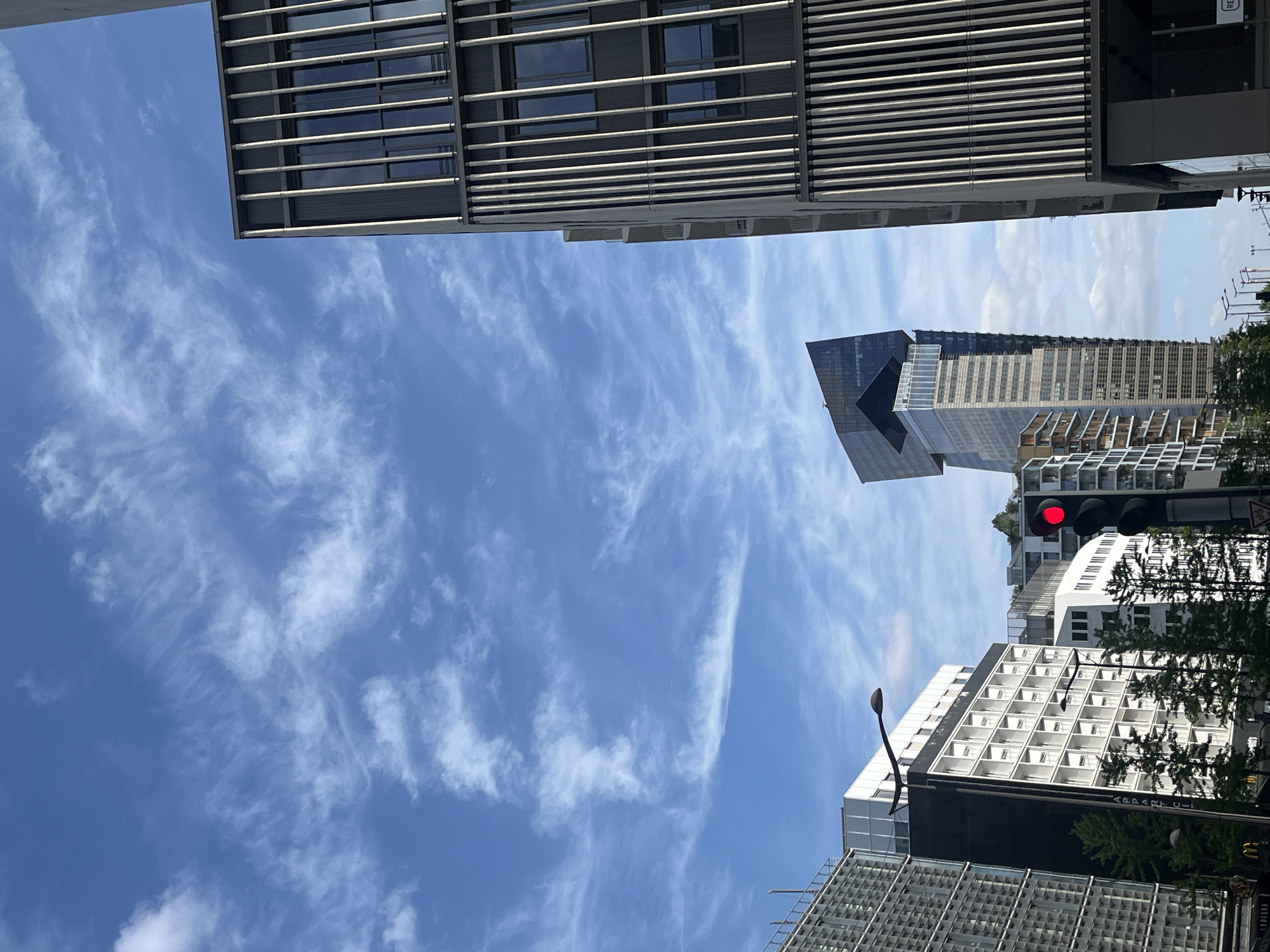 This image presents an upward-looking view of modern urban architecture against a bright blue sky, characteristic of Paris, France. The sky, occupying a significant portion of the frame, is a vibrant blue with scattered, wispy white clouds, suggesting clear weather on a sunny day.
In the upper left quadrant, a large, contemporary building displays a facade with prominent horizontal louvers or fins covering its dark windows. Towards the middle and right, a distinctive, tall skyscraper dominates the view, featuring an angular, dark-colored, possibly glass or metal, top section that gives it a unique silhouette. Below this striking apex, the building transitions to lighter sections with a regular grid pattern of windows and balconies.
Further down on the right side, several other multi-story buildings are visible. One white-faced building exhibits a consistent grid of square windows or balconies. Directly below it, another building features a prominent black horizontal band with white text clearly reading "APART'CITY," indicating a serviced apartment or hotel.
Interspersed among the buildings, some green foliage from trees or bushes is visible, particularly towards the lower right, partially obscuring parts of the structures. A traffic light with a red illuminated signal is visible on the right side, and a slender, dark street lamp or decorative fixture curves upwards in the lower right. No specific human activity is depicted; the scene focuses on the architectural landscape during bright daylight.