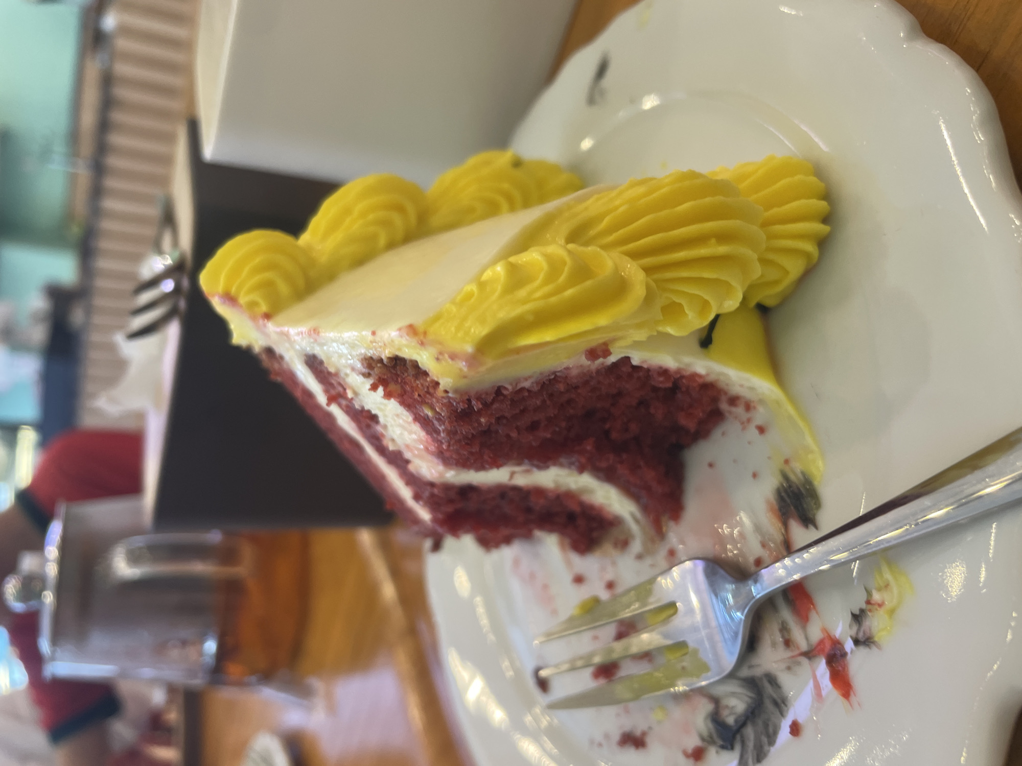 A slice of red velvet cake with yellow frosting sits on a white plate, with a fork resting on the plate. The cake is partially eaten. In the background, a glass teapot and a white mug are visible on a wooden table. A person wearing a red shirt is also partially visible in the background. The scene appears to be in a restaurant or cafe.