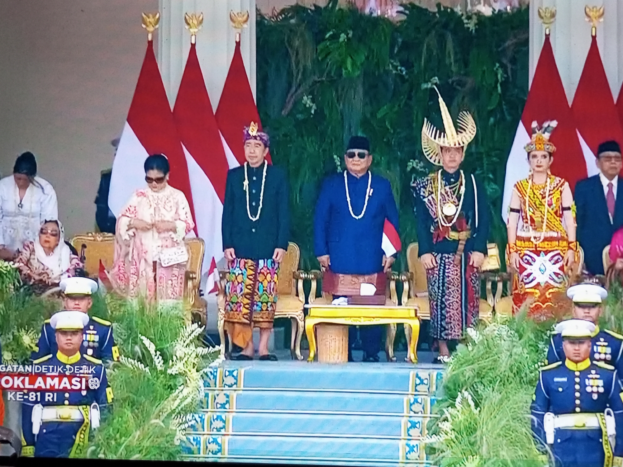 The image shows a formal ceremony taking place on a stage in front of Indonesian flags. In the foreground, two men in dark blue military-style uniforms with white caps stand facing forward. Behind them, an assortment of people are gathered. Several individuals are wearing traditional Indonesian attire, including colorful patterned fabrics and ornate headwear. One person wears a distinctive golden headdress. A woman in the center of the image is wearing sunglasses and a patterned dress. A man in a blue suit and black hat is also present. The backdrop consists of lush green foliage and decorative elements. The stage itself appears to be elevated, with blue stairs adorned with a patterned motif. The overall scene suggests a significant cultural or political event. The text "GATAN DETIK-DETIK PROKLAMASI KE-81 RI" is visible in the lower left corner, indicating the event is related to the 81st anniversary of Indonesia's Proclamation of Independence. The location is specified as Banyudono, Indonesia.