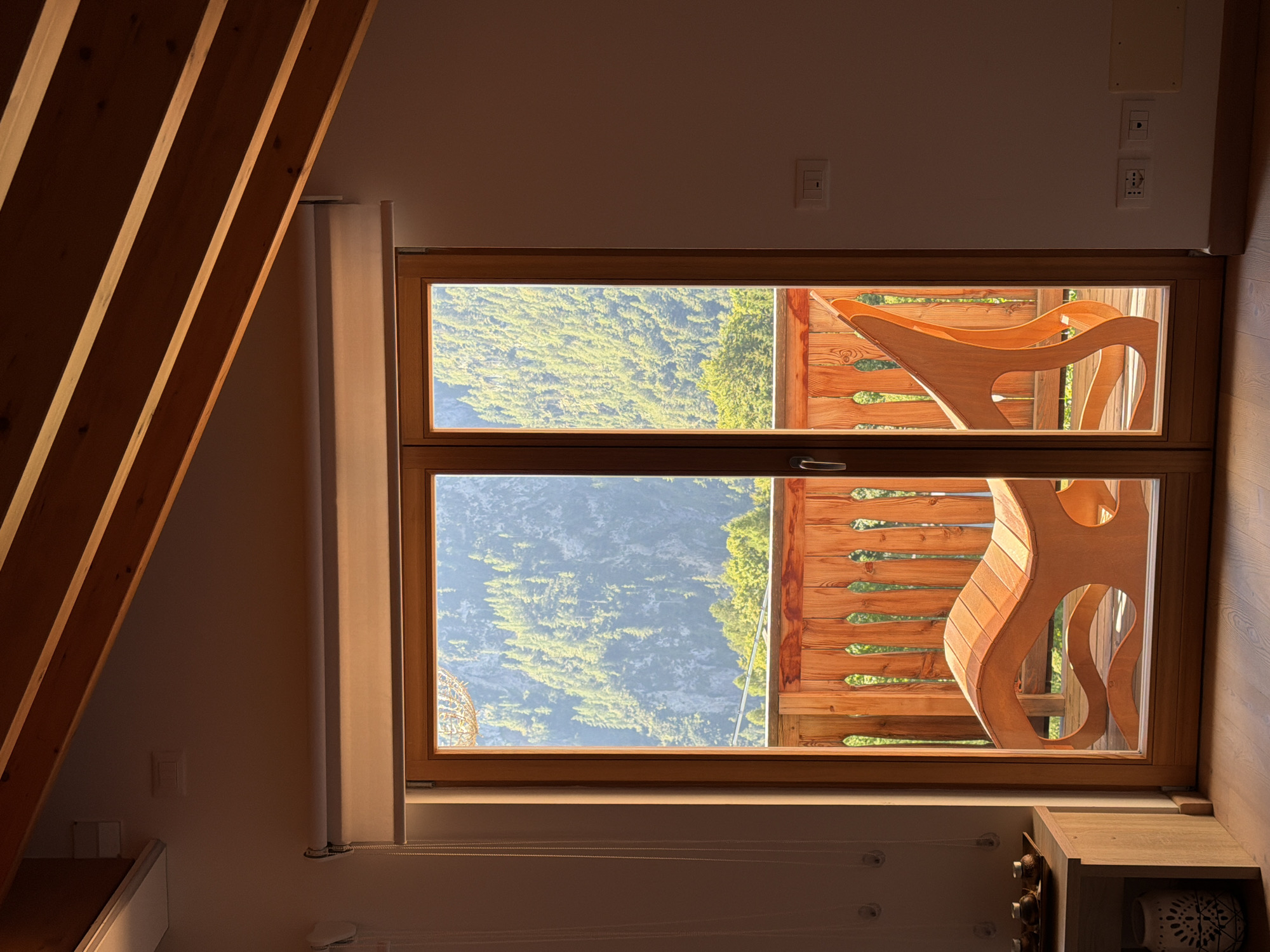 An interior shot focuses on a large wooden-framed glass door leading to a balcony with a wooden railing. Through the door, a sunlit mountain slope covered in dense green trees is visible. Inside, a wooden chaise lounge with a flowing, sculptural design is positioned on the balcony. To the left of the door, a small wooden table holds decorative items, and a decorative vase sits on the floor. The ceiling features exposed wooden beams. The room appears to be in a mountainous region, likely a hotel or private residence. Given the context provided, this scene is located in Sauris / Zahre, Italy.