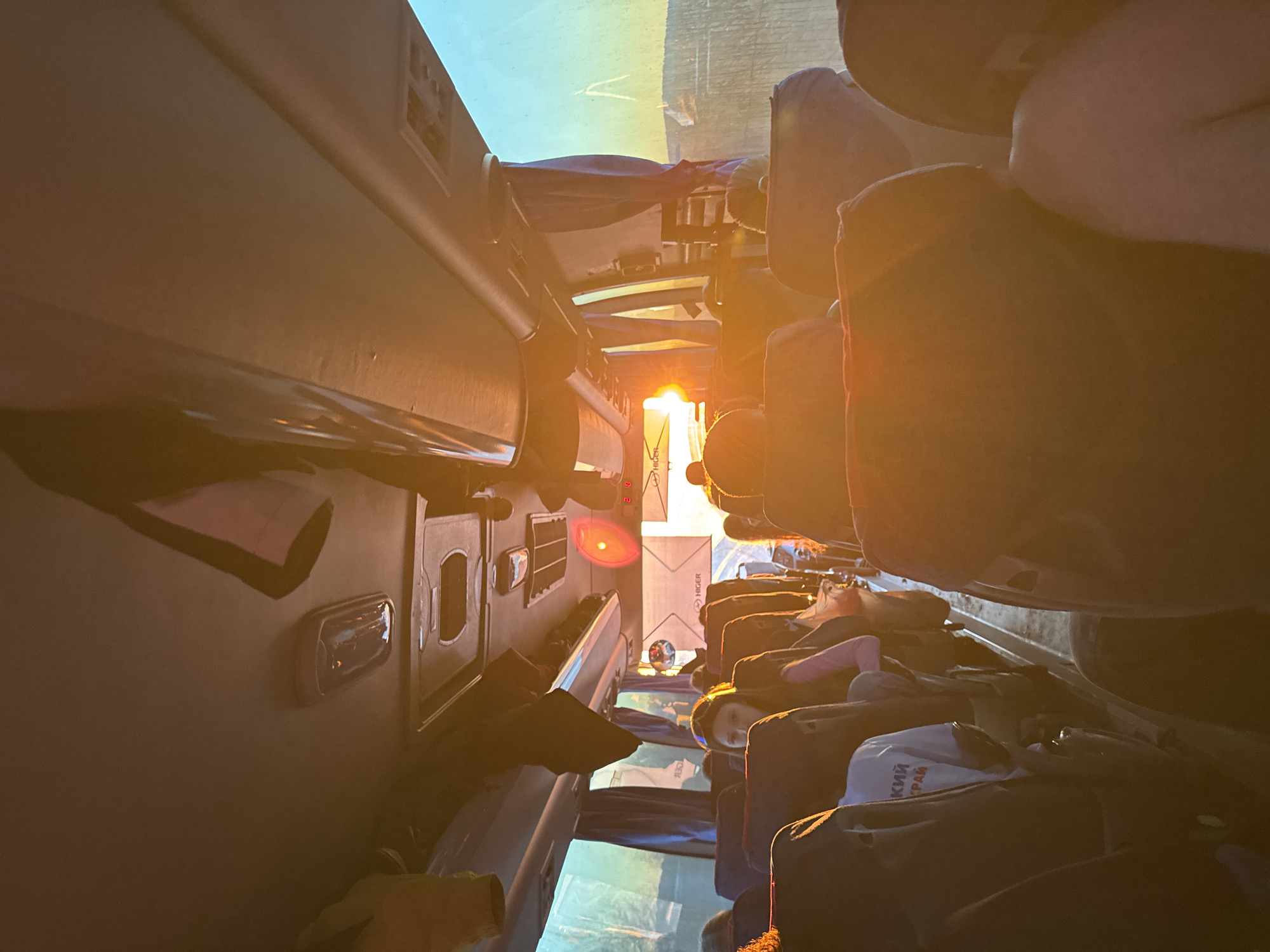 The image depicts the interior of a bus, likely traveling during sunrise or sunset, based on the golden light. Passengers are seated in rows, with their belongings stowed around them. Text in Cyrillic is visible on a bag. A label with "HIGER" can be seen near the windshield. The precise location in Ustinovo, Russia cannot be determined from this image alone.