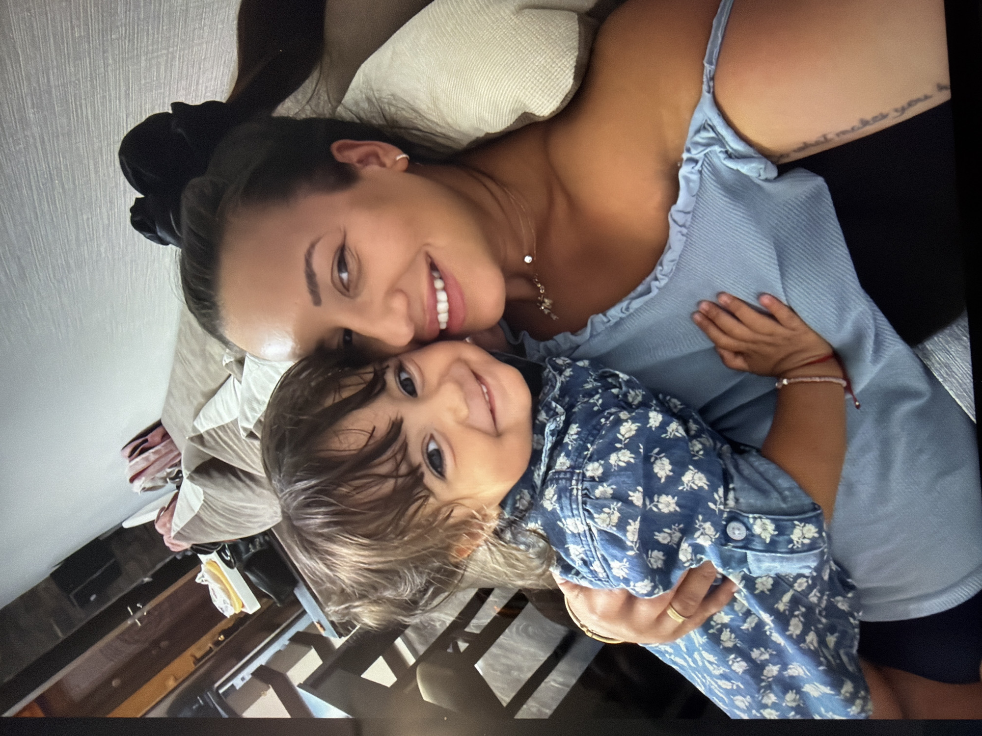 A light-skinned woman with dark hair in a bun is lying down, holding a young child with brown hair and eyes. The woman is wearing a light blue top and a necklace. The child is wearing a blue floral shirt. Both are smiling at the camera. There is a tattoo visible on the woman's upper arm. The background includes a pillow, bedding, and furniture. The location in Burgas, Bulgaria is unknown.