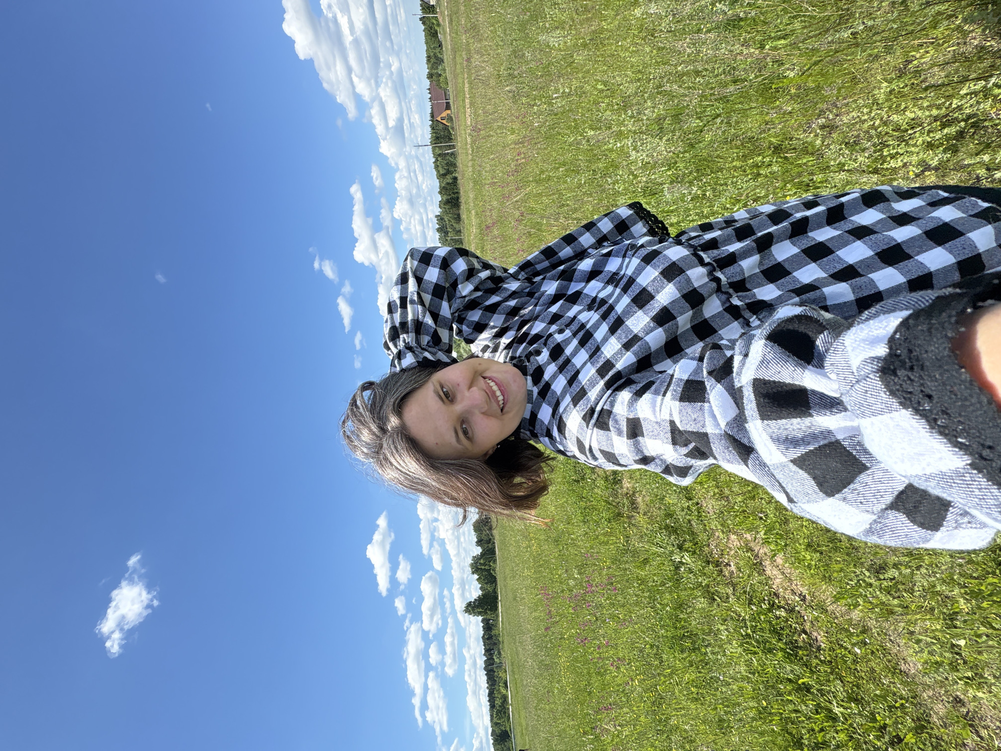 A young woman with shoulder-length graying hair stands in a grassy field under a bright blue sky dotted with fluffy white clouds. She is wearing a black and white checkered dress and has her right arm raised behind her head, with her left arm extending toward the camera. The background shows a line of trees in the distance, with a few houses visible among them. The field is filled with tall green grass and scattered wildflowers. The image appears to be a selfie taken outdoors on a sunny day.