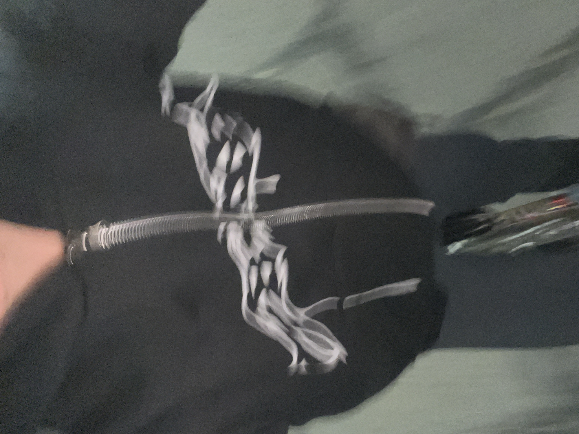 A blurry image shows a black garment, possibly a jacket or sweater, with a white graphic design. The design appears stylized, perhaps a skull or abstract figure, with sharp, angular features. A metal zipper runs across the garment. The background is a mix of light and dark tones. Given the blur, it's impossible to pinpoint the exact location in Olivos, Argentina.