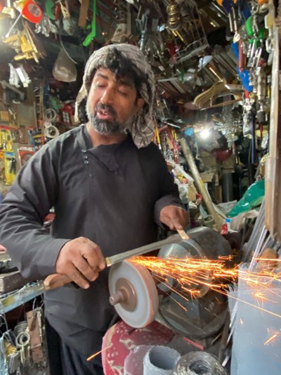 The image depicts a man in Kandahar, Afghanistan, sharpening a knife. The man is wearing a dark-colored garment and a head covering. He is holding a knife to a spinning grinding wheel, producing sparks. The background is cluttered, displaying various tools and hardware items, suggesting the location is a workshop or small retail space. The environment appears modest and utilitarian, reflecting the conditions and resources of a local business in Kandahar.