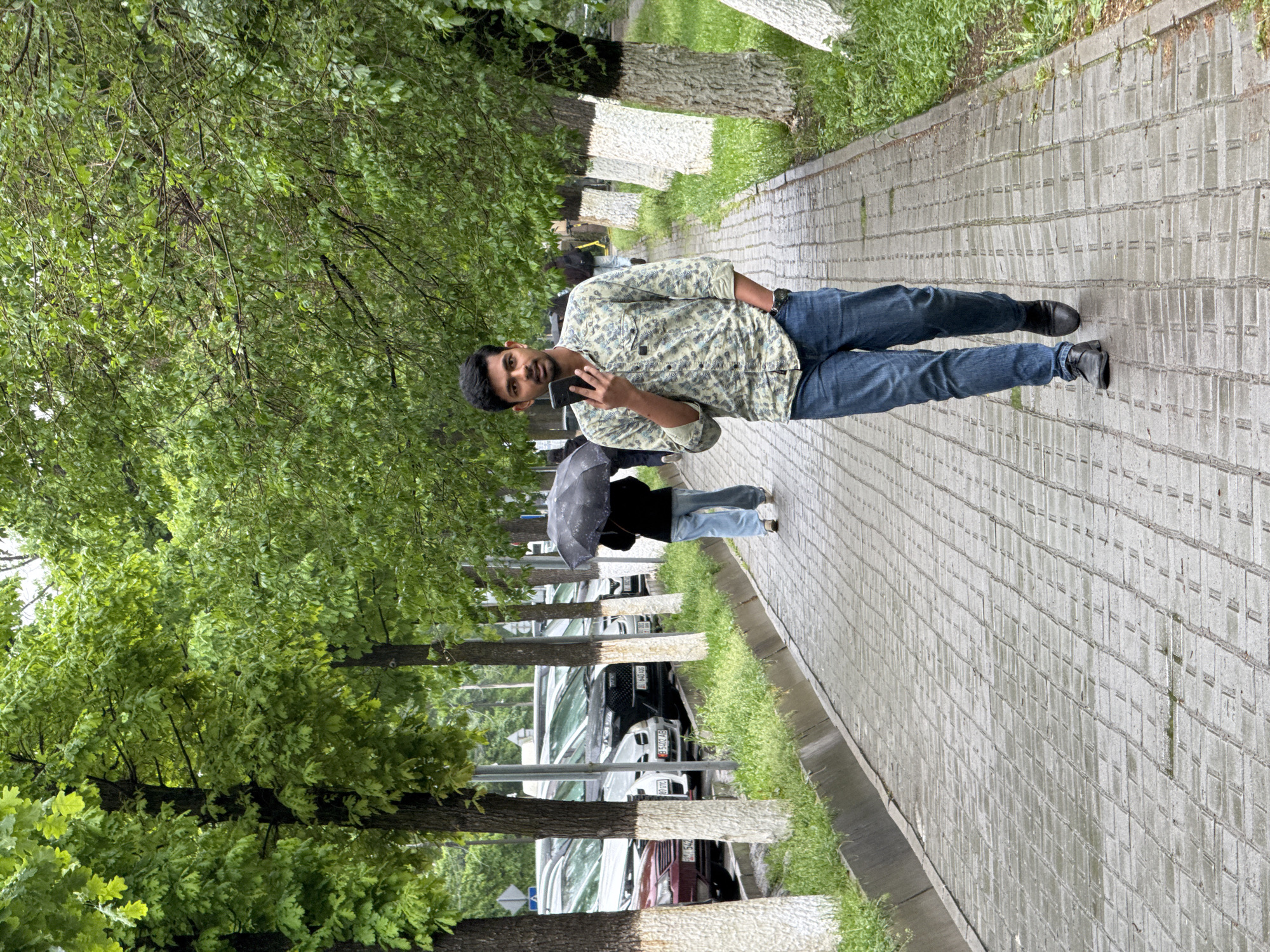 This outdoor photograph, taken in Bishkek City, Kyrgyzstan, captures a street scene after recent rainfall. The image features a young man in the foreground, standing on a paved sidewalk and looking directly at the camera while holding a black smartphone in his left hand. He is dressed in a light-colored, patterned long-sleeve shirt, blue jeans, and dark shoes.
Behind him, further down the path, another individual is partially visible, walking away and holding a black umbrella, reinforcing the impression of wet weather. The grey rectangular paving stones of the sidewalk appear damp.
The path is flanked on one side by a row of tall, leafy green trees with dense foliage, suggesting late spring or summer. A notable detail is that the lower sections of the tree trunks are distinctively whitewashed. Beyond these trees, a line of parked cars is visible, with some Kyrgyz license plates showing Cyrillic characters and numbers (e.g., "B 3404 AB" and "02 KG"). On the other side of the sidewalk, a narrow strip of green grass separates the path from what appears to be a curb or another section of the road.
The lighting suggests an overcast daytime, consistent with the rainy conditions. The overall ambiance is fresh and verdant, reflecting a typical day in the city during the warmer months.