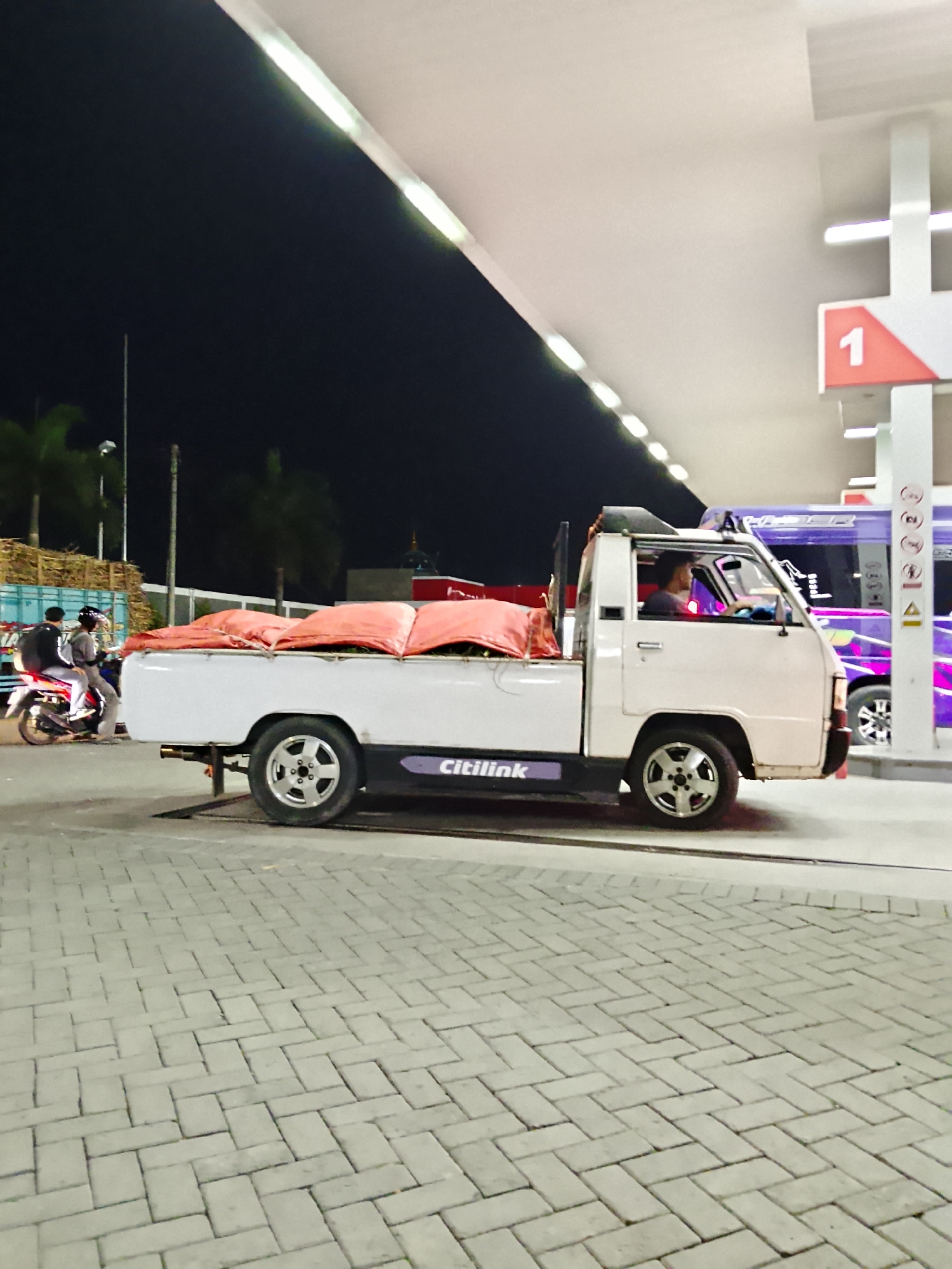 At a gas station in Kasiman, Indonesia, a white pickup truck is parked under a covered canopy. The truck bed is covered with bright orange tarps, suggesting it is loaded with goods. A man is visible in the driver's seat. In the background, a purple and pink bus is parked. To the left of the pickup truck, two individuals are riding a motorcycle. The ground is paved with gray interlocking bricks. The scene is illuminated by artificial lighting from the gas station canopy and the surrounding area, indicating it is nighttime.