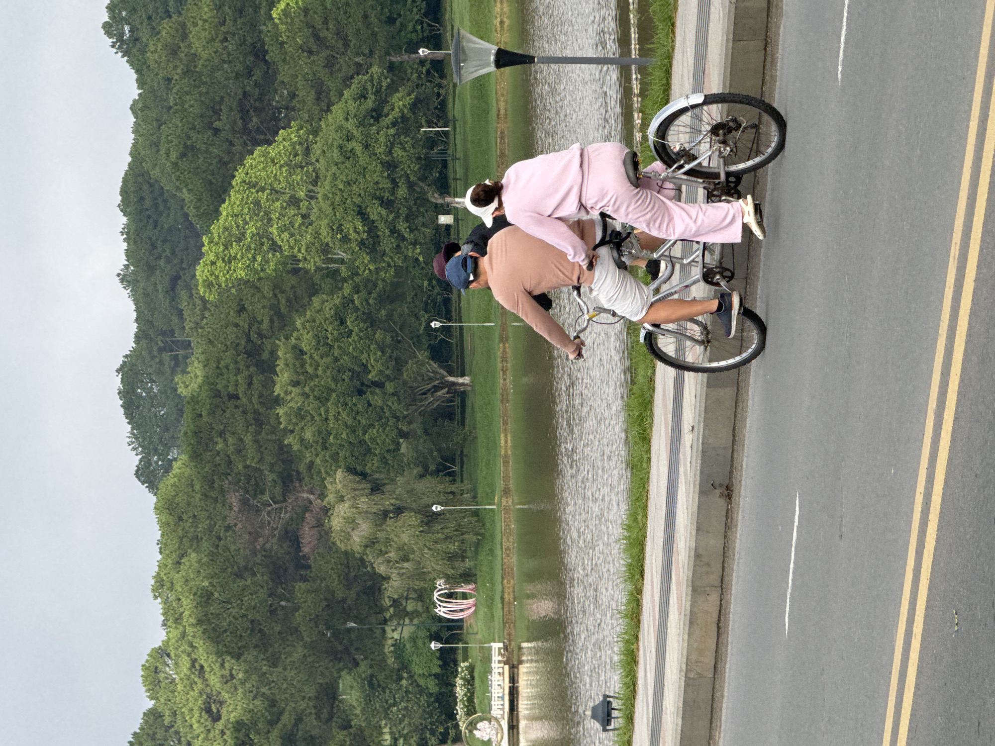 Two individuals, a man and a woman, are riding a tandem bicycle on a paved road beside a body of water. The man, positioned in front, wears a tan long-sleeved shirt and light-colored shorts. The woman, seated behind him, is dressed in a pink tracksuit and a white cap. They are both facing away from the camera. The tandem bicycle is silver with two sets of pedals and wheels. The road is gray asphalt with yellow lines marking the lanes. Behind the road is a concrete sidewalk and a strip of grass. Beyond the grass is a lake, with a dock and some decorative sculptures on the left side. Further in the background, a dense forest of green trees covers rolling hills under an overcast sky. This scene is identifiable as Xuan Huong Lake in Da Lat, Vietnam, due to the distinctive landscape and the presence of a tandem bicycle, which are common tourist attractions there.