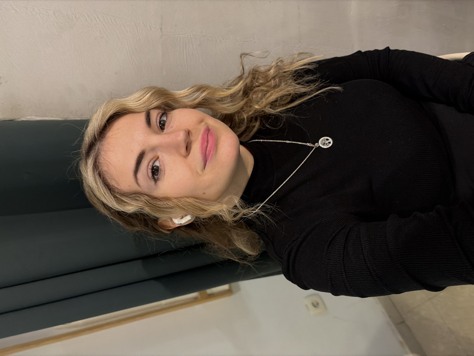 A woman with curly blonde hair and fair skin smiles at the camera. She wears a black turtleneck sweater and a silver necklace with a round pendant. A white earbud is visible in her left ear. The background is a wall with a dark green panel on the left. A light switch is visible near the bottom right corner of the image.