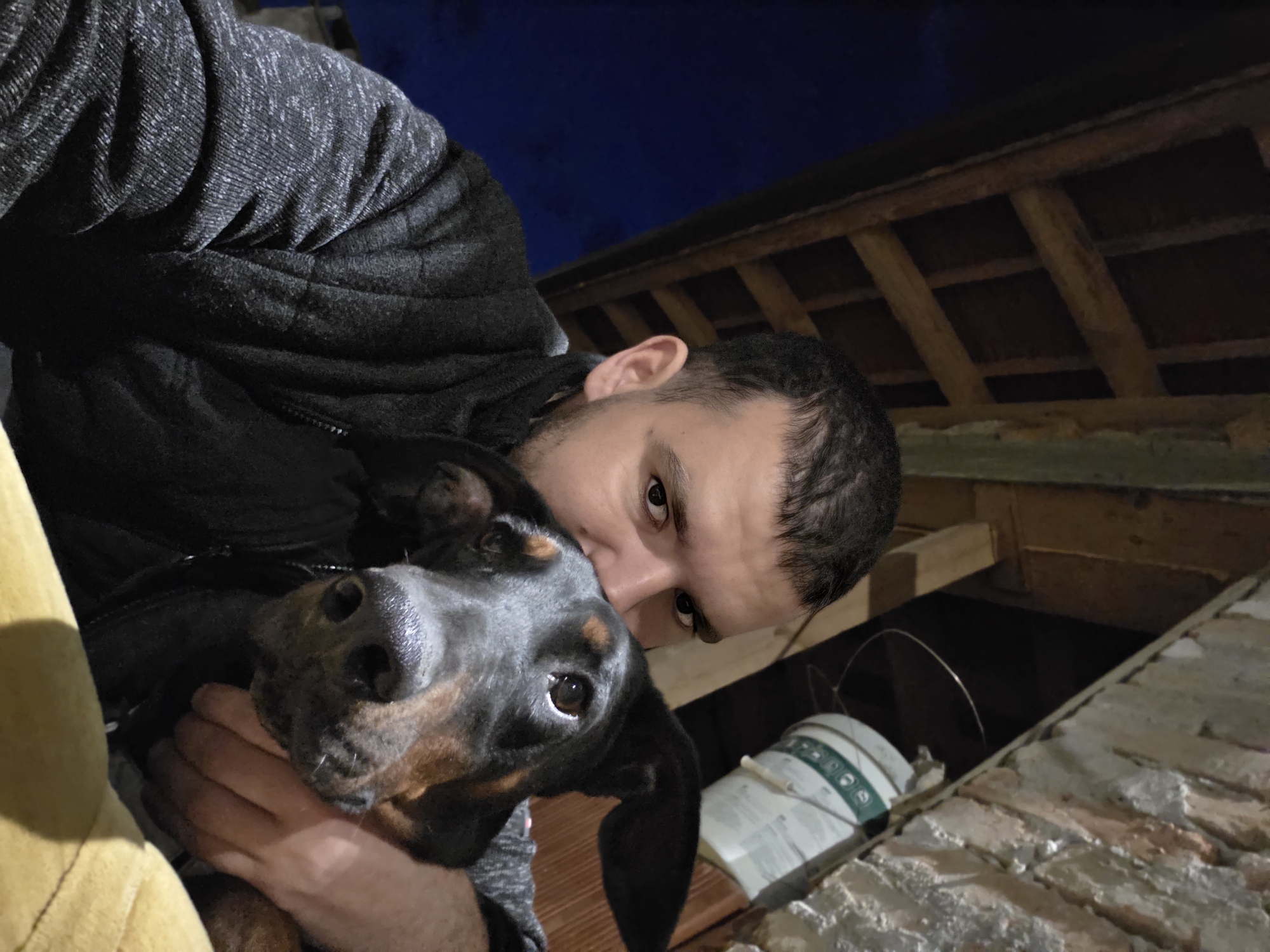 The image displays an adult male holding a black and tan dog in a selfie-style composition. The male, positioned on the left, has short dark hair and stubble, wearing a grey textured long-sleeved shirt beneath a dark grey puffer vest. He looks directly at the camera. The dog, possibly a hound mix, is on the right, with its head resting against the man's chest/shoulder. The man's right arm embraces the dog. The background exhibits exposed wooden beams forming a ceiling structure in the upper left, with a dark blue area further left. To the upper right, exposed brickwork and a white bucket with green labeling are visible. A yellow fabric surface occupies the lower right foreground. The scene is artificially lit, indicative of a low-light indoor or covered outdoor environment. Location: Unknown, Unknown.