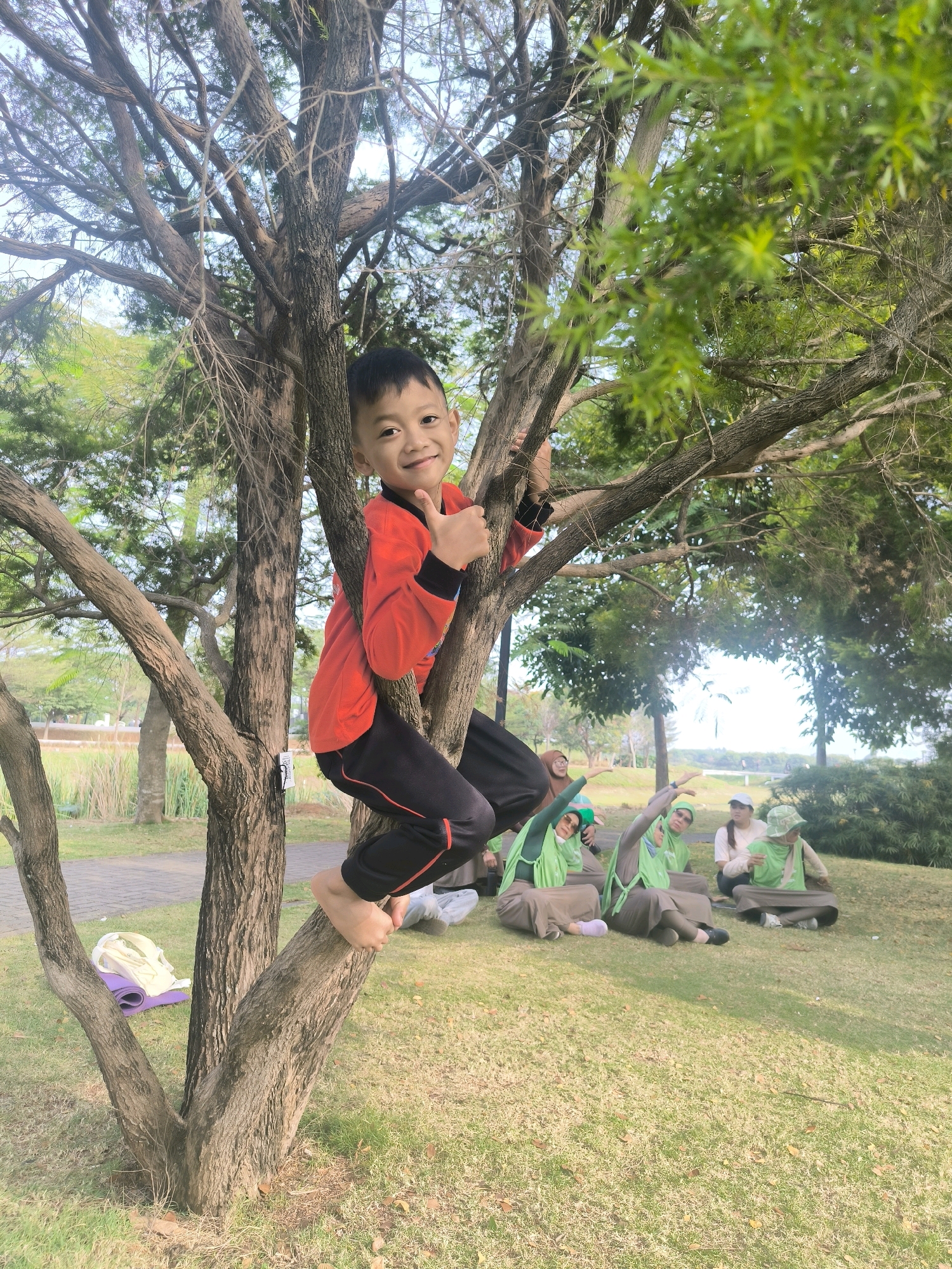 A young boy in a red long-sleeved shirt and black pants with red trim is climbing a tree and giving a thumbs-up. He is positioned on a thick branch, with his body turned to the right. His feet are bare. In the background, several people are sitting on the grass, some wearing green hijabs. The scene is outdoors in a grassy area with trees and a body of water visible in the distance. The location is the Bandung City Botanical Garden, Bandung, Indonesia.