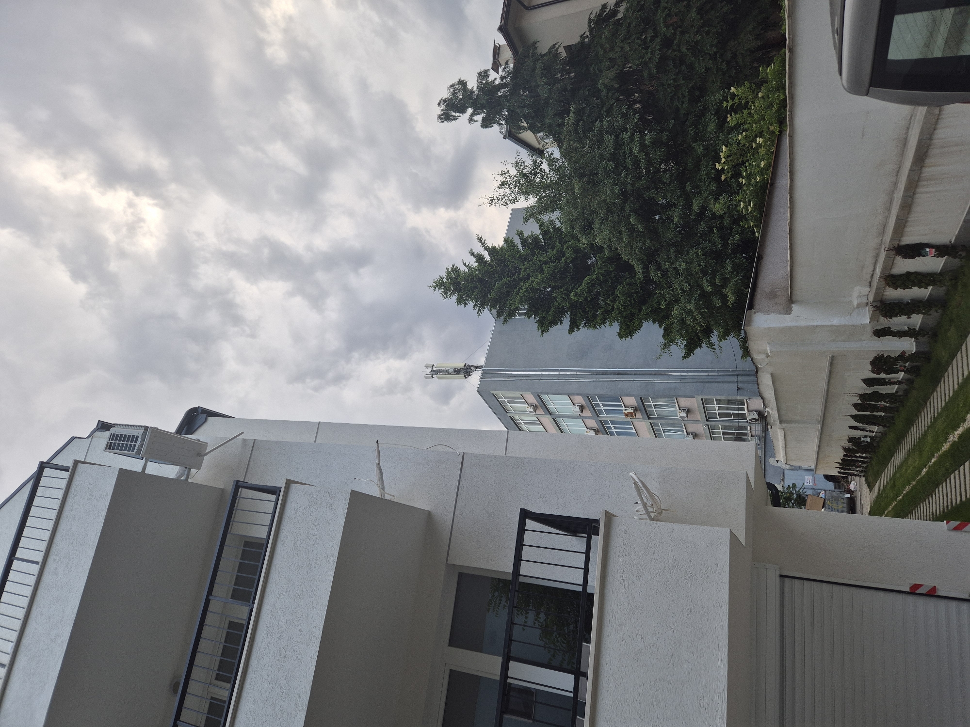 The image captures an urban or suburban landscape under an overcast sky. The scene is dominated by architectural structures, primarily focusing on a modern, white building with stark, angular lines and black metal railings on its windows and balconies. This building has a textured, possibly stucco, facade. An outdoor unit, likely an air conditioner, is visible on one of its upper levels.
In the background, a taller, older grey building with multiple rectangular windows and external air conditioning units can be seen. On its roof, a cellular antenna tower or similar communication equipment is mounted. Lush, tall dark green trees are interspersed between the buildings, providing a natural element to the structured environment.
To the right, a silver car is partially visible. Below it, a neatly maintained area features striped patterns of green grass alternating with concrete paths or dividers, suggestive of a driveway or garden pathway. A large, vertically paneled garage door in a light color occupies the lower part of the scene, and nearby, a white concrete wall features red and white striped barriers.
The sky is cloudy and grey, with some brighter patches indicating diffused light, suggesting either an overcast day, early morning, or late afternoon. No people or discernible activities are taking place; the scene appears still and quiet. There is no visible text or specific location cues to identify the exact geographic setting.