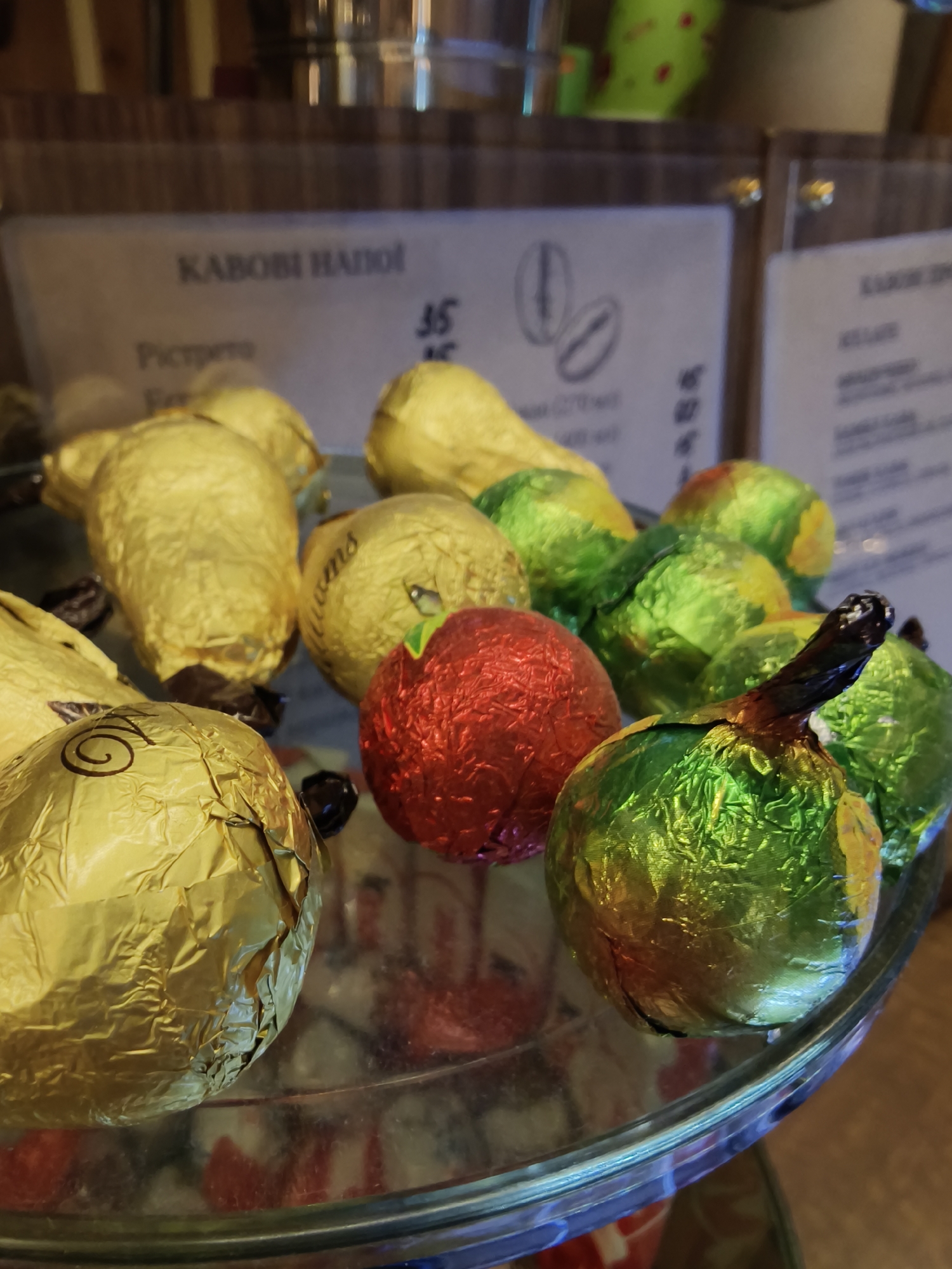 A high-angle, close-up shot shows a glass tray of foil-wrapped candies, in a business establishment identified by signage in the background as "KABOBI HAHOI," a cafe or shop in Kryvyi Rih, Ukraine. The candies appear to be shaped like fruits (apples, pears) and are wrapped in shiny foils of gold, green, and red. The foreground candies are clearly visible and the background is blurred. The focus is on the assortment of candies, reflecting ambient light on the surface of the glass tray.