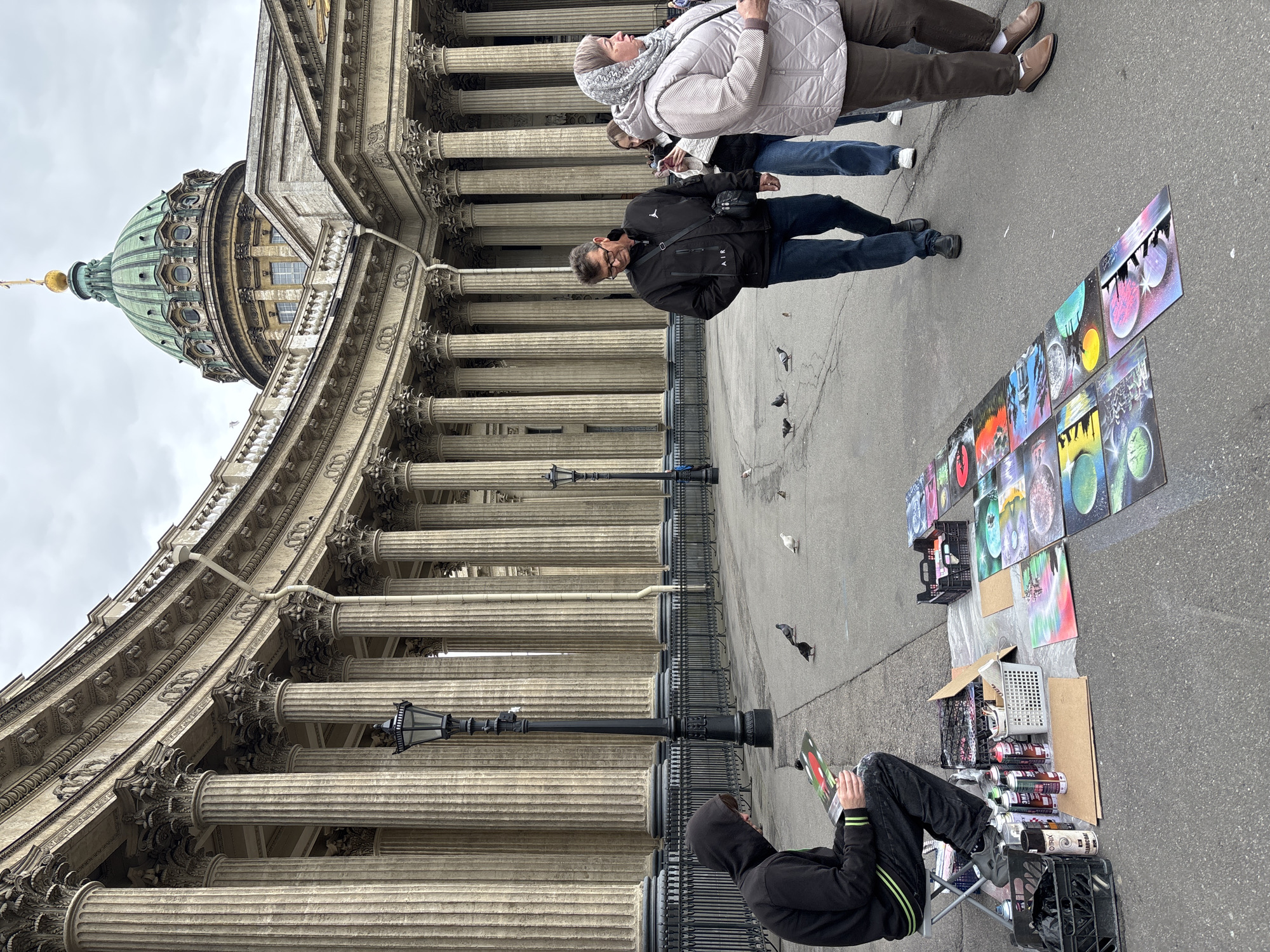 The image captures a scene outside the Kazan Cathedral in Saint Petersburg, Russia. The architectural details of the cathedral, including its colonnade and green dome with a golden spire, are prominent. A street artist is sitting on a folding chair, surrounded by spray paint cans and milk crates filled with supplies. Several paintings are displayed on the ground beside them. Tourists are walking past the artist along the sidewalk, and a few pigeons are scattered nearby on the pavement.
