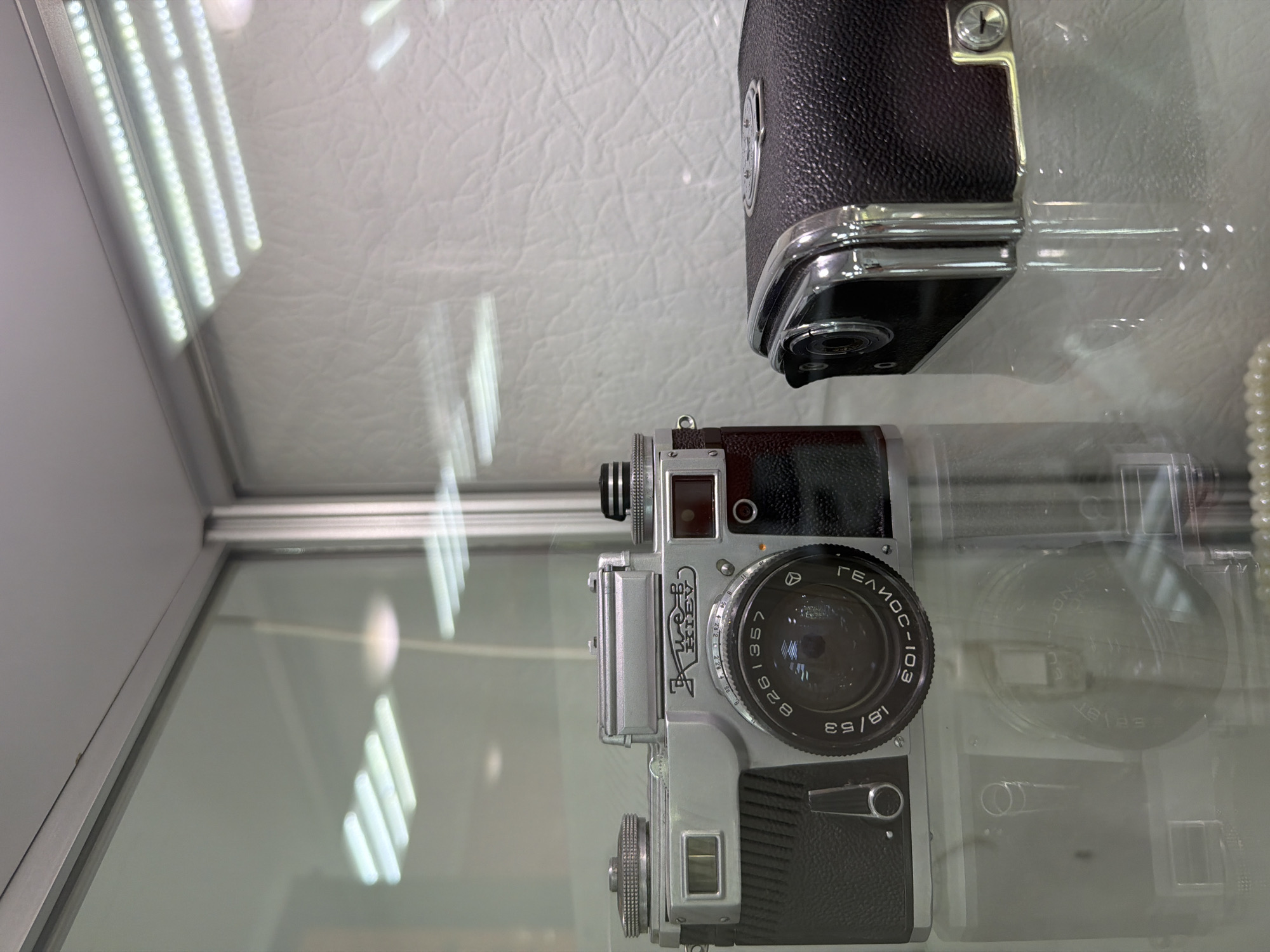 Two vintage cameras are displayed in a glass-enclosed cabinet. The camera on the left is silver with a black textured grip and a prominent lens. The camera on the right is predominantly black with chrome accents. The cabinet's interior features a white, textured background and is illuminated by overhead lights. The image is a close-up shot, focusing on the details of the cameras. The scene suggests a museum or exhibition setting. There is no information in the image to determine its location in Tobolsk, Russia.