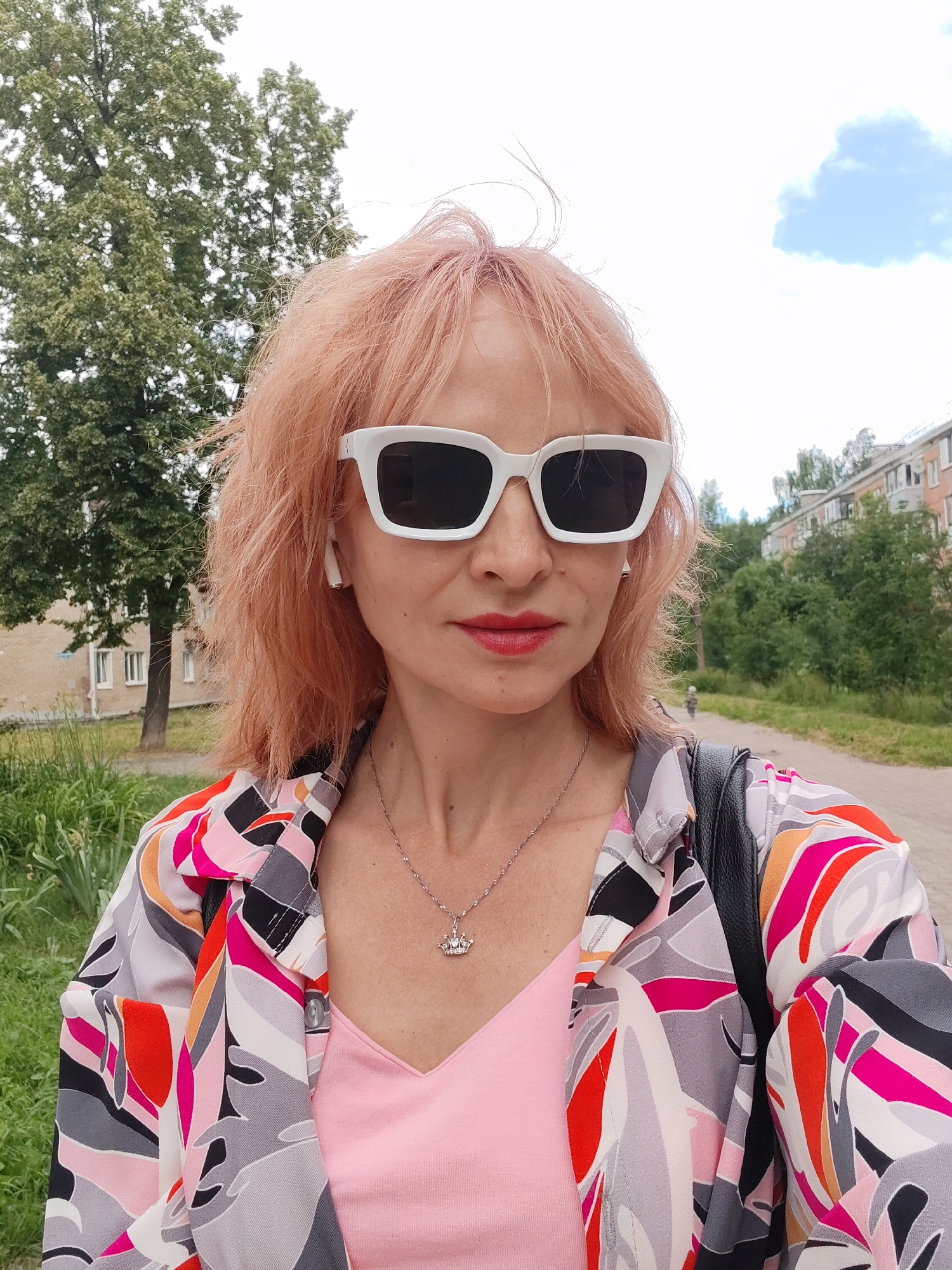 A woman with peach-colored hair, wearing large white sunglasses, a pink top, and a colorful patterned jacket, stands outside. She has earbuds in her ears and a silver necklace with a crown pendant. Behind her, a tree and a building can be seen. The setting appears to be a street in the city of Chaikovsky, Russia, indicated by the style of the buildings and the general environment.