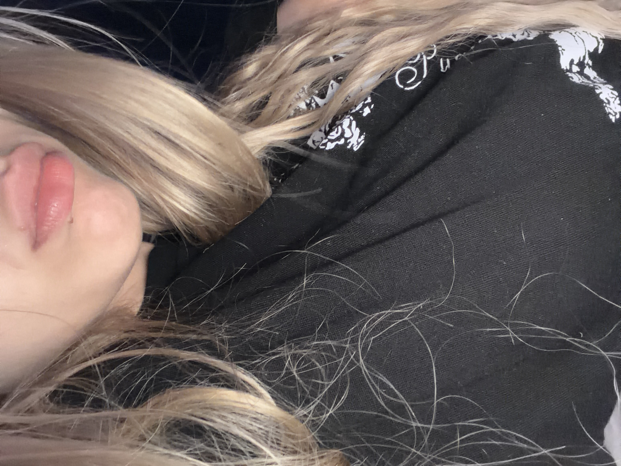 A close-up, head-and-shoulders shot focuses on the lips and part of the face of a person with blonde hair. The individual is wearing a black top with a white graphic design. The background is dark and out of focus. The image appears to be taken indoors, possibly in a dimly lit room. There are no discernible landmarks to indicate a specific city or location.