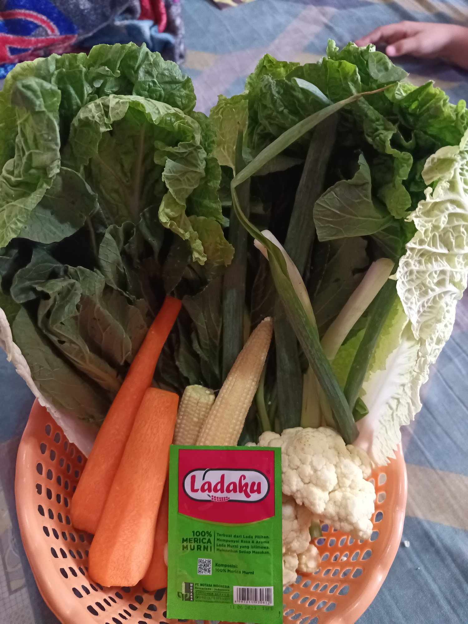 In South Jakarta, Indonesia, an overhead shot showcases a basket filled with fresh vegetables. The basket, made of orange plastic with a perforated design, contains a variety of produce. Prominently featured are two peeled carrots, their bright orange color contrasting with the green of the vegetables. Several baby corn cobs are visible, their pale yellow kernels clustered together. A head of cauliflower, with its distinct white florets, sits towards the right side of the basket. Green leafy vegetables, including what appears to be Napa cabbage and possibly some leafy greens like bok choy, fill a significant portion of the basket. A packet of "Ladaku" brand merica murni (pure pepper) with a green background and red lettering is placed in the foreground, partially obscuring the vegetables. The basket rests on a surface that appears to be a blue and white checkered cloth, possibly a tablecloth or mat. In the upper portion of the image, parts of a person's hand and some colorful fabric, potentially clothing or a blanket, are visible, suggesting a domestic setting. The lighting is bright, illuminating the textures and colors of the vegetables.