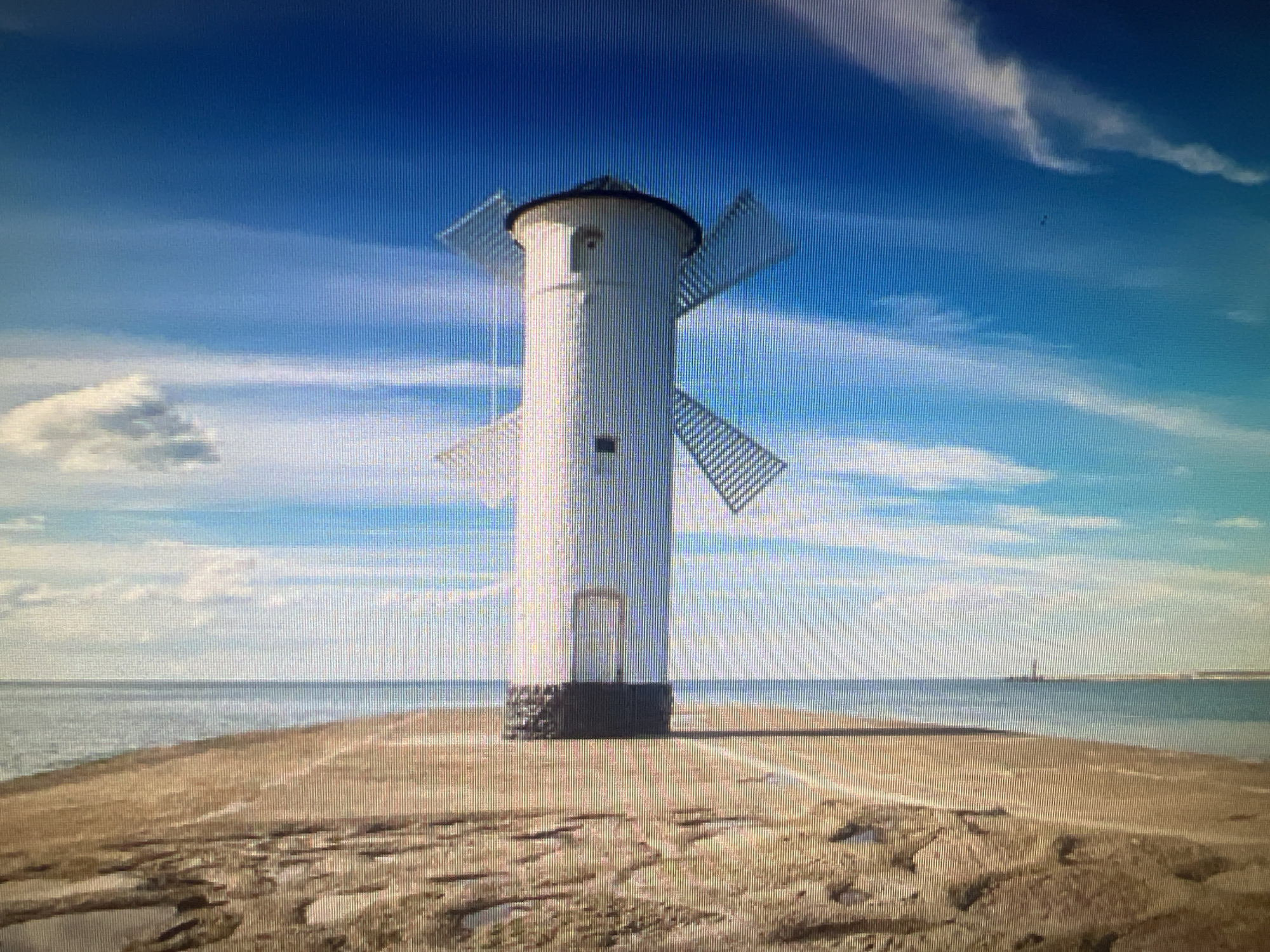 The image displays a lighthouse with windmill-like sails, situated on a sandy pier extending into the sea. The sky is a vibrant blue with scattered clouds, and the sea is calm and reflects the sky. The lighthouse is white with a dark roof and has a door and a small window on its cylindrical body. The sails are angled, suggesting they are not currently rotating. The ground is a mixture of sand and what appears to be rocks or breakwater elements. In the far distance, another lighthouse or structure is visible on the horizon. The scene is bathed in sunlight, creating a bright and clear atmosphere. There are no people visible in the image. This specific lighthouse, known as Stawa Młyny, is located in Świnoujście, Poland, not Cheb, Czechia.