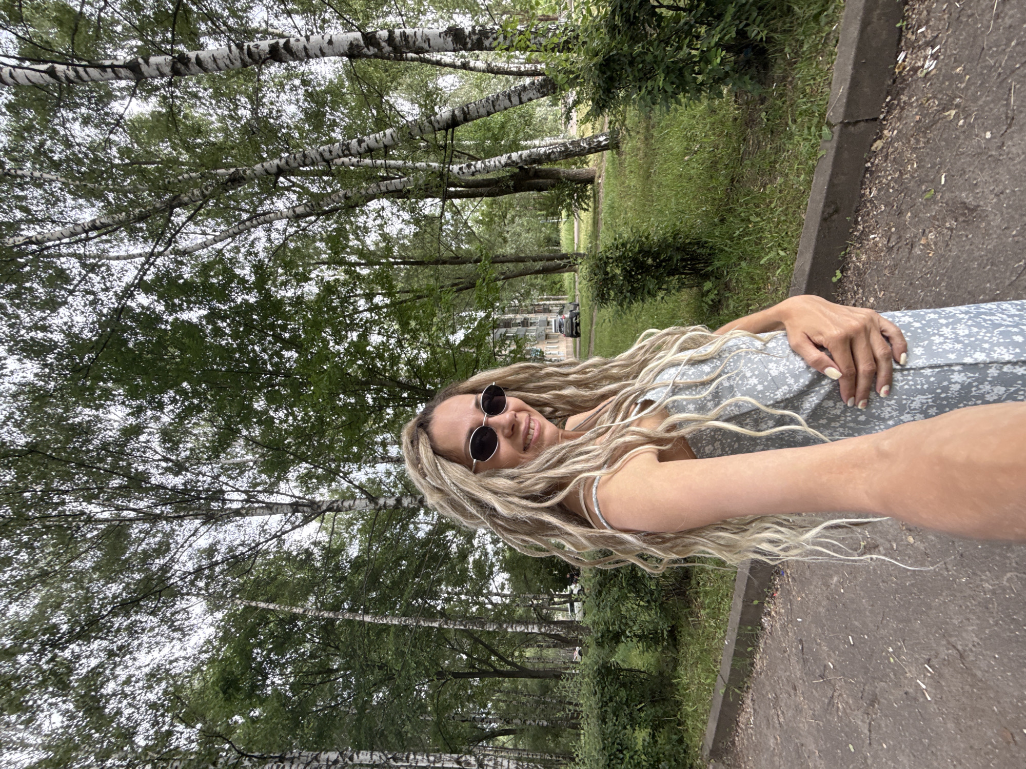 A young woman with blonde dreadlocks and sunglasses smiles at the camera, holding her arm out as if taking a selfie. She is wearing a light blue and white floral dress and stands on a paved walkway in front of a line of birch trees. The background features a grassy area with more trees and a building in the distance. The scene appears to be a park or residential area.