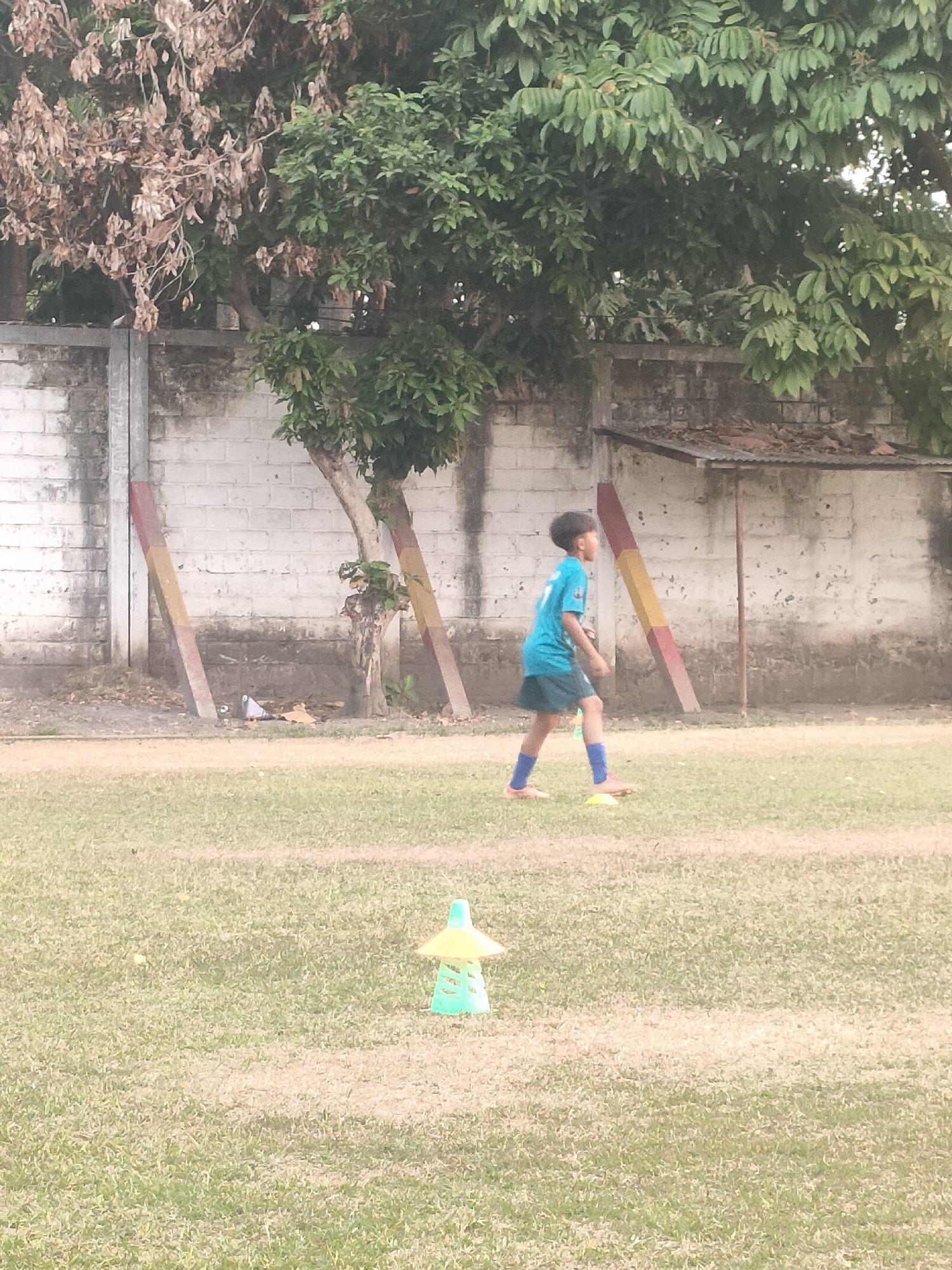 A young boy in a blue soccer jersey and green shorts, with blue socks and pink shoes, is running across a grassy field. He is the only person visible in the image. The field has a few green and yellow cones placed on it, suggesting a practice or training session. In the background, there is a weathered white brick wall with some graffiti or staining. Adjacent to the wall are some trees, one of which has dried, brown leaves, while others are lush and green. There are also some wooden posts with red and yellow diagonal stripes leaning against the wall. A simple open-sided structure with a corrugated metal roof is visible near the wall. The overall scene suggests an outdoor sports area in a semi-urban or rural setting. This scene is likely located in Banguntapan, Indonesia, given the context.