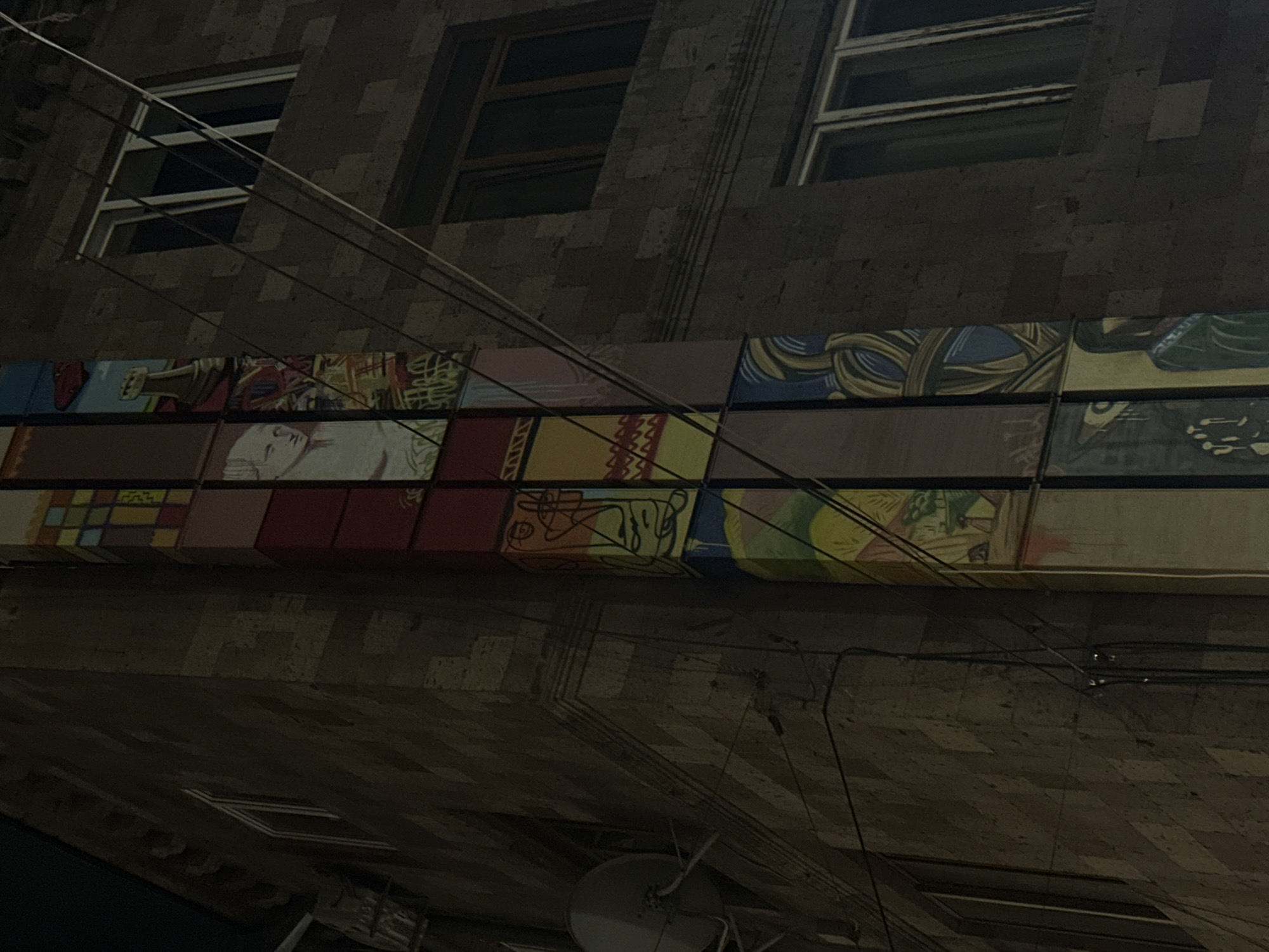 The image shows the exterior of a building in Yerevan, Armenia. The building appears to be constructed of stone or brick, with a row of windows visible. A distinctive feature is the band of colorful, graphic panels running horizontally across the facade. Electrical wires are prominent, crossing the frame and obscuring parts of the building. Below the painted panels, the ceiling/roof features a satellite dish and additional wires. The lighting suggests that the photo was taken at night or in low-light conditions.