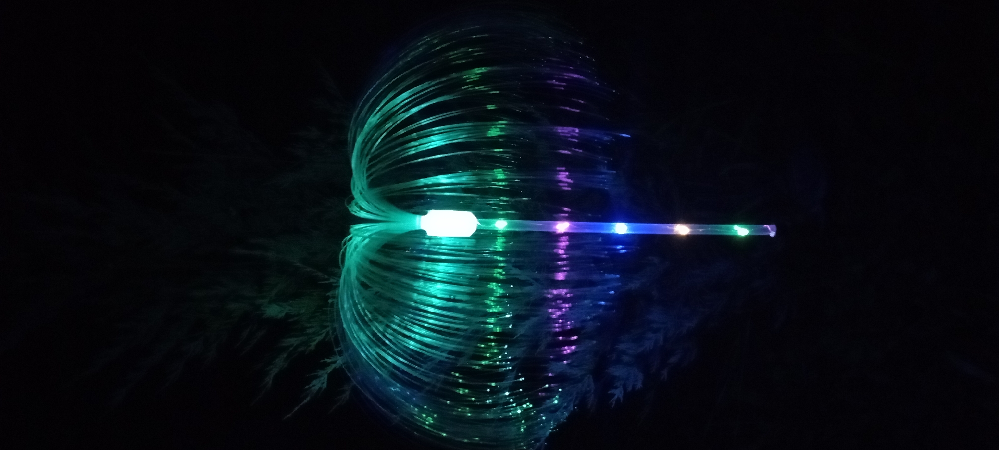 In the city of Radisne, Ukraine, a solar-powered fiber optic lamp is illuminated at night. The lamp, resembling a jellyfish or a dandelion, is designed with multiple fiber optic strands that emit a gradient of green and purple light. These strands fan out from a central cylindrical core, which also features embedded LEDs. The base of the lamp is a thin pole, also illuminated by LEDs. The background is dark, indicating nighttime, with a hint of greenery from a nearby plant or tree visible at the top. The ground is not clearly visible, but the overall impression is of an outdoor garden or yard setting.