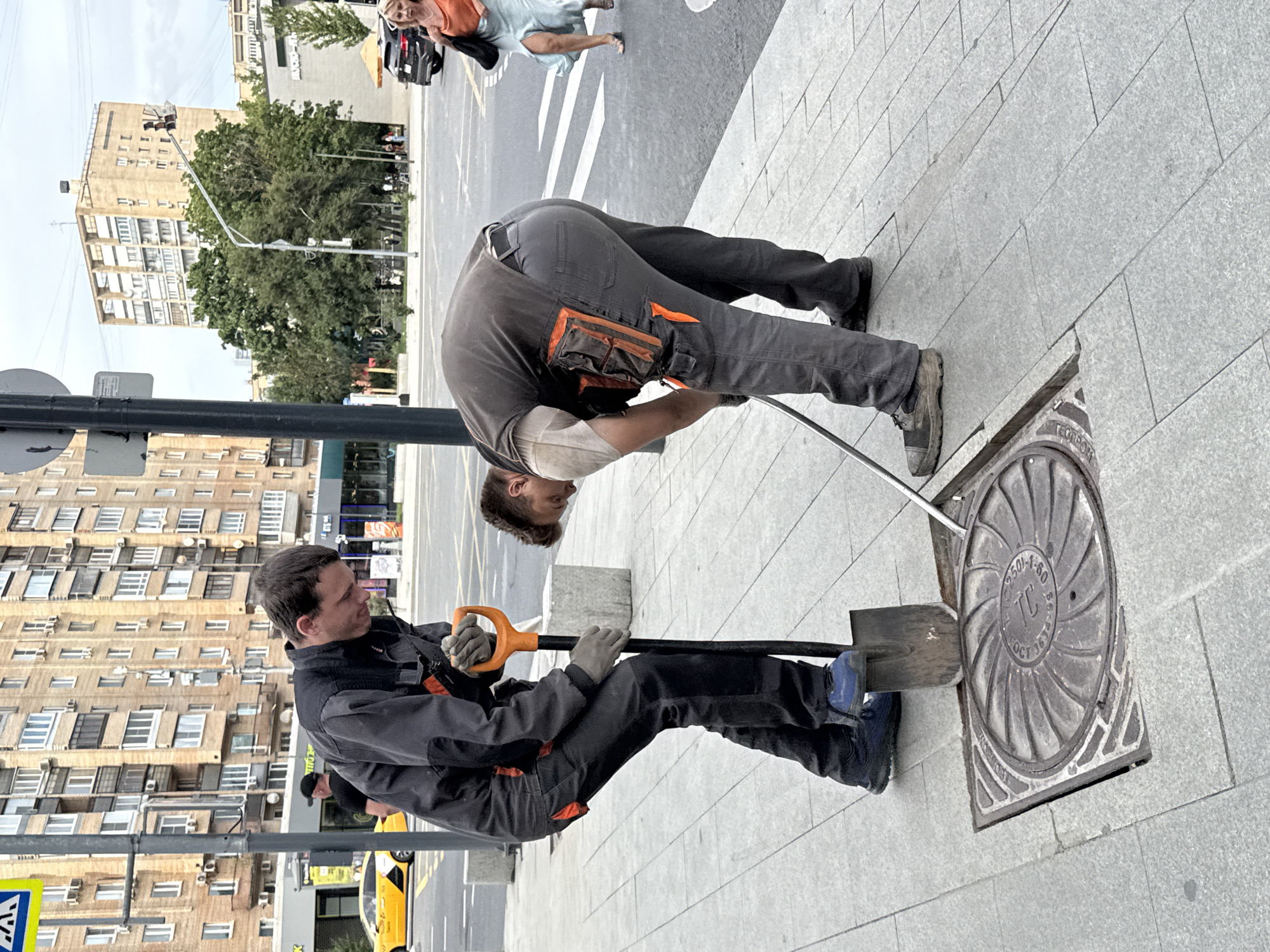 Two male construction workers are seen in the image, engaged in what appears to be maintenance work on a manhole cover in Moscow, Russia. The manhole cover is circular with intricate patterns and text, and it is partially lifted from its housing. One worker, wearing a dark gray jumpsuit with orange accents, is using a shovel with an orange handle to pry at the manhole cover. He is wearing gray work gloves and blue work boots. The other worker, dressed in a gray vest over a light-colored shirt and dark gray pants with orange utility pockets, is bending down and appears to be using a long, thin metal rod to assist in lifting the manhole cover. He is also wearing dark work shoes. Both workers are on a paved sidewalk made of large, light gray square tiles.
In the background, a street with yellow and white markings is visible, with a yellow taxi car driving on it. Several multi-story apartment buildings with many windows and balconies line the street. A traffic light pole is also present. Further in the background, a woman in an orange top and a light blue skirt is walking away from the camera, and a black SUV is parked on the side of the road. Trees with lush green foliage are interspersed between the buildings and along the street. The sky is overcast. The overall scene suggests an urban environment where public infrastructure maintenance is taking place.