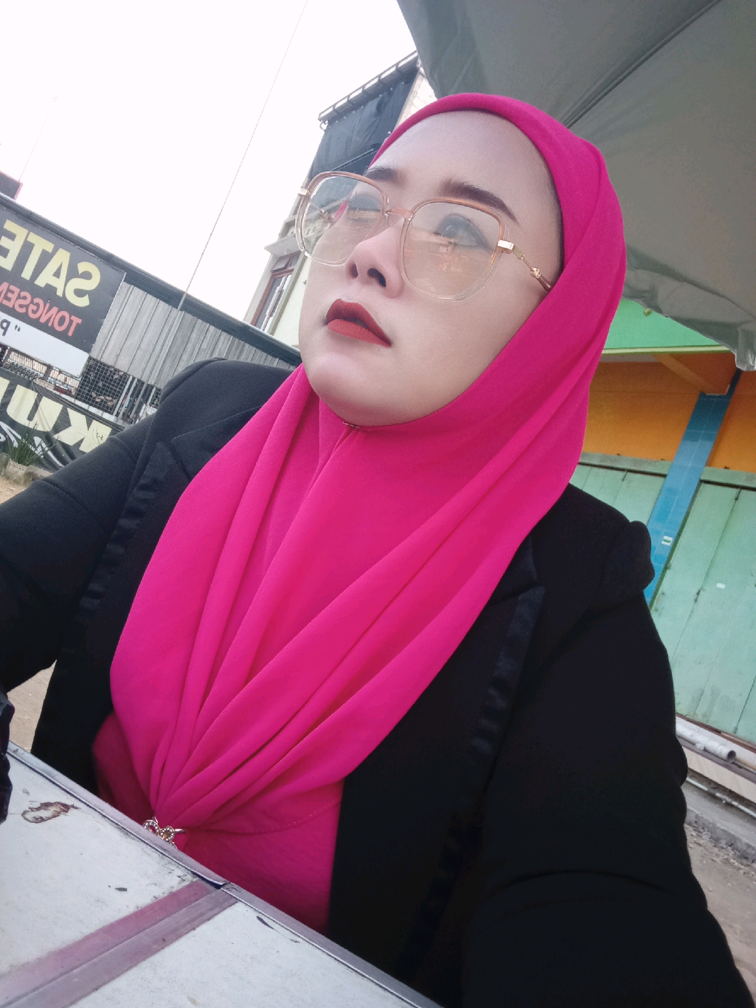 A woman wearing a bright pink hijab and a black jacket is seated at a table, looking upwards and to her left. She is wearing large, clear-rimmed glasses and red lipstick. A decorative brooch is visible on her hijab, fastening it at the chest. In the background, a sign with yellow lettering is partially visible, appearing to read "SALE" and "TONGSEM". The environment suggests an outdoor setting, possibly in Simo, Indonesia, with a light-colored, possibly overcast sky. The woman is positioned in the foreground, with the background elements slightly out of focus.