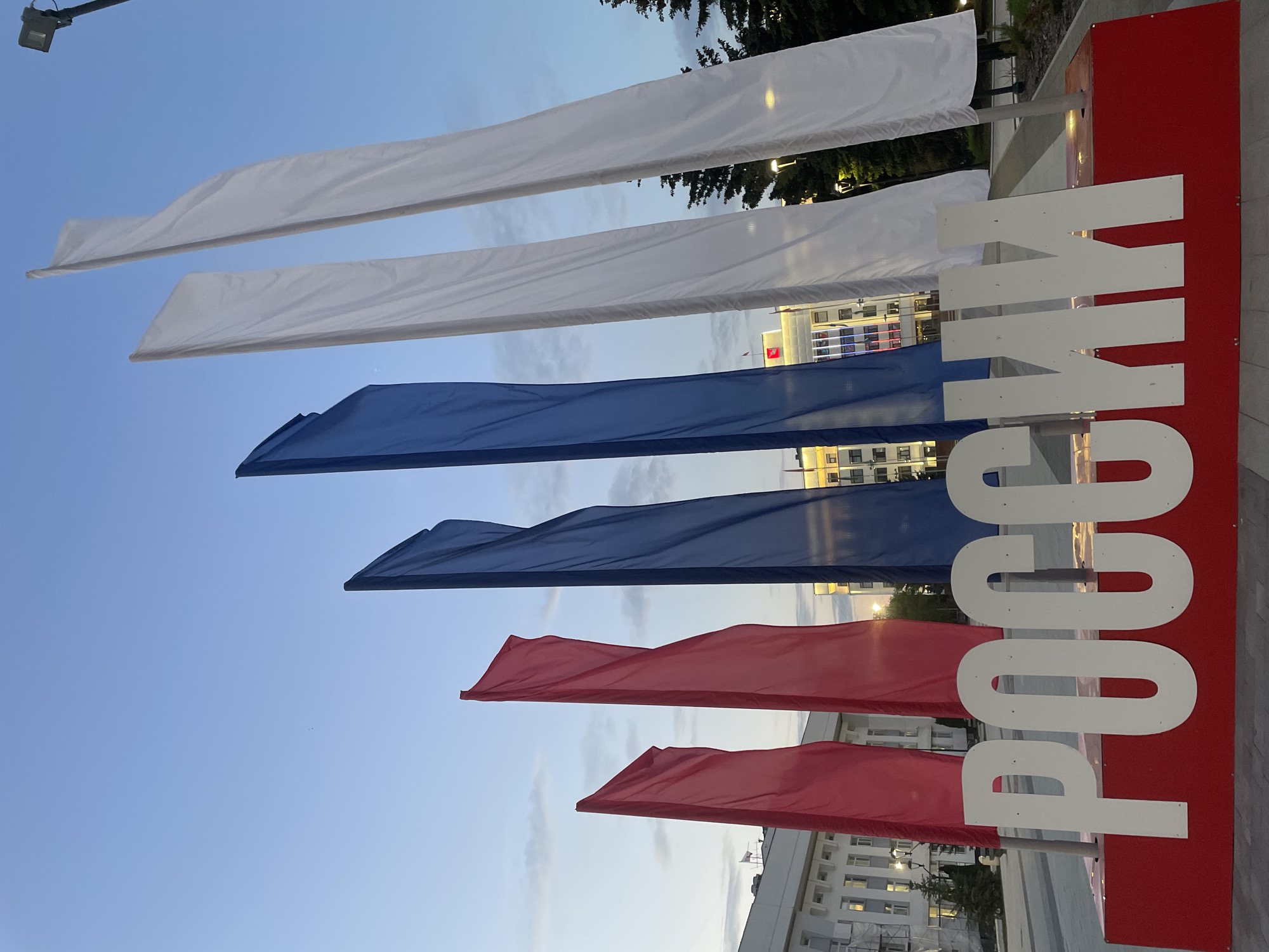 The image displays a patriotic display with the colors of the Russian flag: white, blue, and red. Several large rectangular flags, or banners, are suspended vertically against a clear sky, transitioning from white at the top, to blue in the middle, and red at the bottom. To the right of the flags, a large, white, three-dimensional inscription reads "РОССИЯ" (Russia). The inscription is mounted on a red rectangular base.
In the background, buildings can be seen, suggesting an urban or public setting. The lighting indicates it is either early morning or late evening, with the sky a soft blue and some clouds present. The overall impression is one of national pride and a public celebration or commemoration. The scene is set outdoors, likely in a public square or street, given the presence of buildings and pavement. There are no people visible in the image. The flags appear to be made of fabric and are gently waving, suggesting a slight breeze. The lighting from the buildings in the background hints that some artificial lights are on, further supporting the time of day being either dawn or dusk.