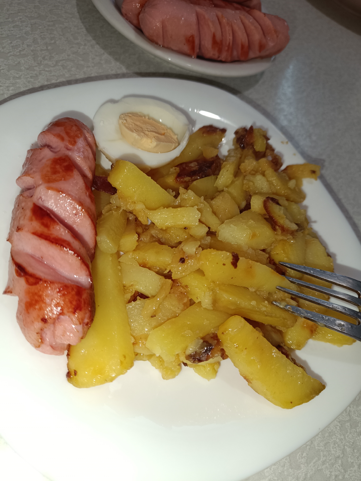 The image shows a plate of food consisting of a grilled sausage, a hard-boiled egg cut in half, and fried potatoes. A fork is resting on the potatoes. In the background, a partial view of another plate with sliced sausages is visible. The setting appears to be a table with a patterned surface. While Tobolsk, Russia is requested in the prompt, this location cannot be determined from the visual information in this image alone. The image does not contain people.