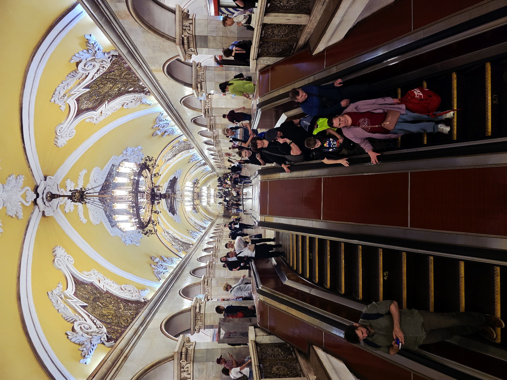 The image depicts the interior of a Moscow metro station, likely Komsomolskaya-Koltsevaya, known for its baroque architecture.
The main focus is the escalator area, with people riding down. Some people are standing and riding the escalator in a standard way. A few people are lying prone on the moving staircase, an unusual and potentially dangerous action. A man at the bottom of the escalator is also lying down, seemingly engrossed with a mobile phone.
The environment is characterized by a high, arched ceiling, painted in yellow and adorned with ornate plasterwork and chandeliers. The station features walls decorated with arched openings and detailed bas-reliefs.