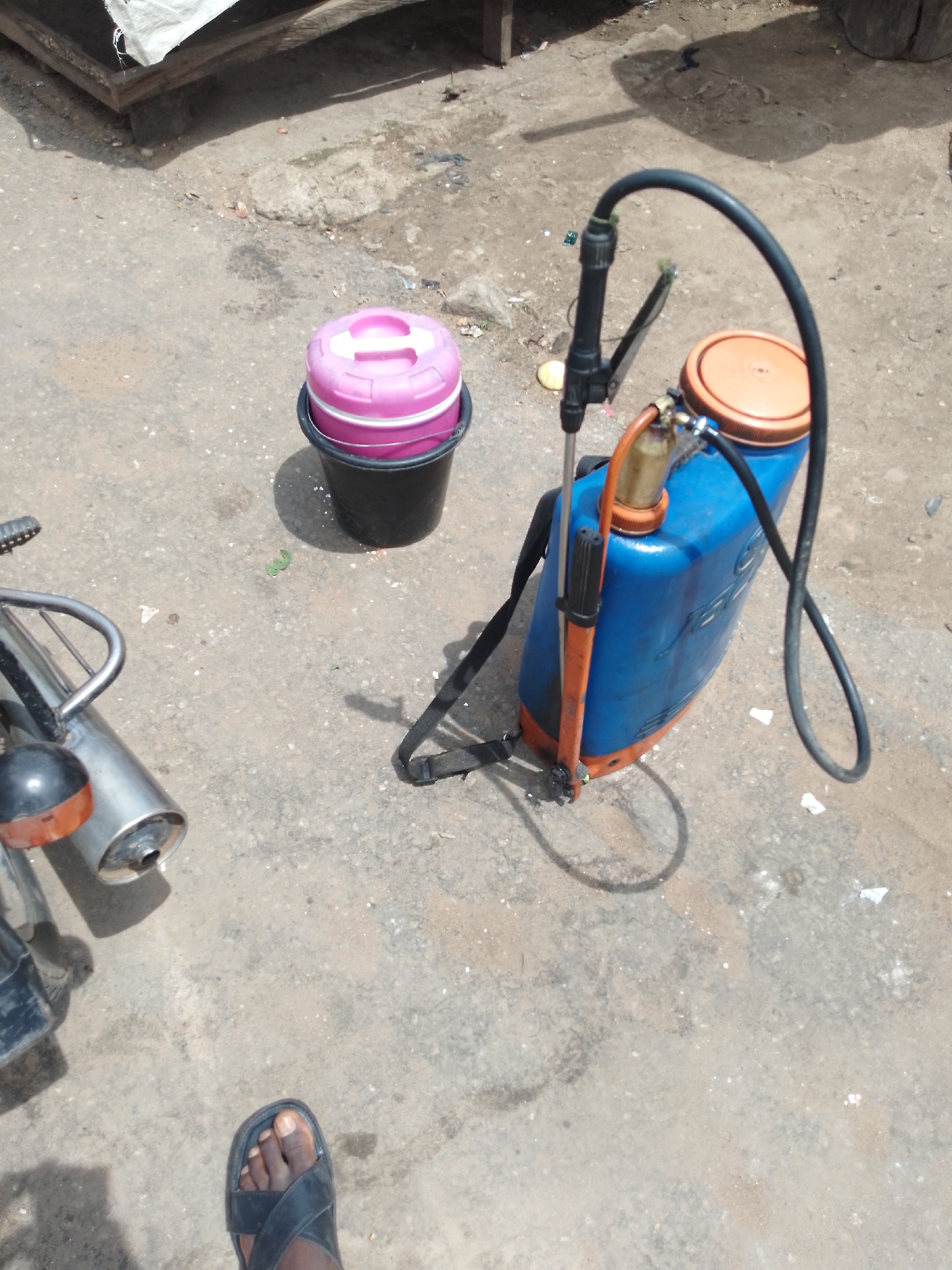 The image captures an outdoor scene on a dusty, concrete surface in Serti, Nigeria. In the foreground, a person's left foot wearing a black sandal is visible. To the left, a portion of a motorcycle is present, including its exhaust pipe and part of the frame. A pink-lidded bucket sits in the middle ground. Behind the bucket, a blue and orange backpack sprayer is the central subject, with its black hose extending upwards. In the background, a wooden structure with white material draped over it is partially visible. The ground is littered with small debris.