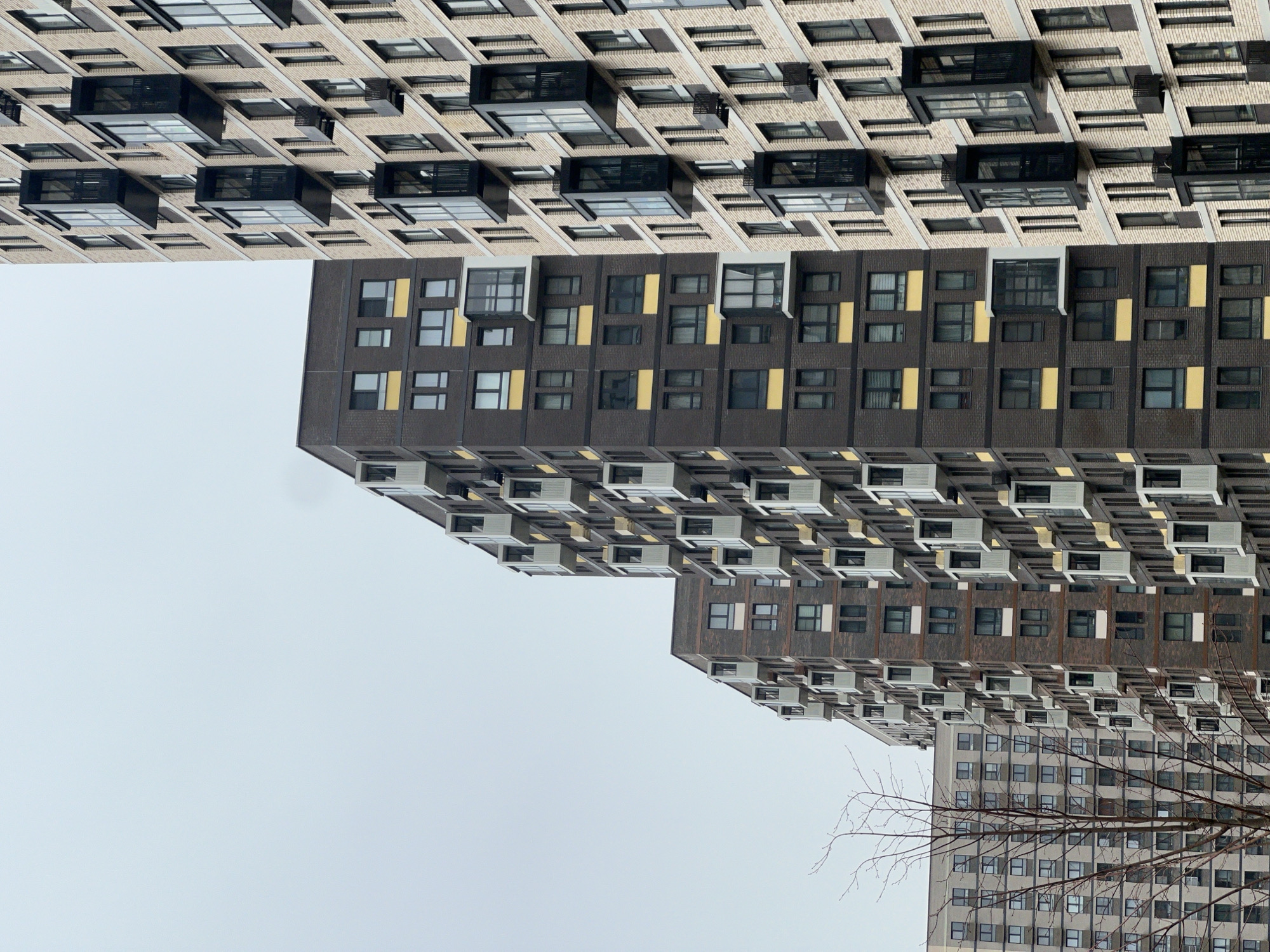 The image depicts a low-angle view of several modern apartment buildings against a gray sky. The buildings are located in Khimki, Russia. The architecture of the buildings is characterized by rectangular shapes, repeated window patterns, and varying color schemes. One building has a light-colored facade with dark window frames and protruding balcony structures. The building beneath has a darker facade with alternating sections of dark and light-colored windows. Below, another building has a facade with many white boxes and windows. In the lower right, a tree without leaves is visible. The photo is taken from a perspective that emphasizes the height and density of the urban environment.