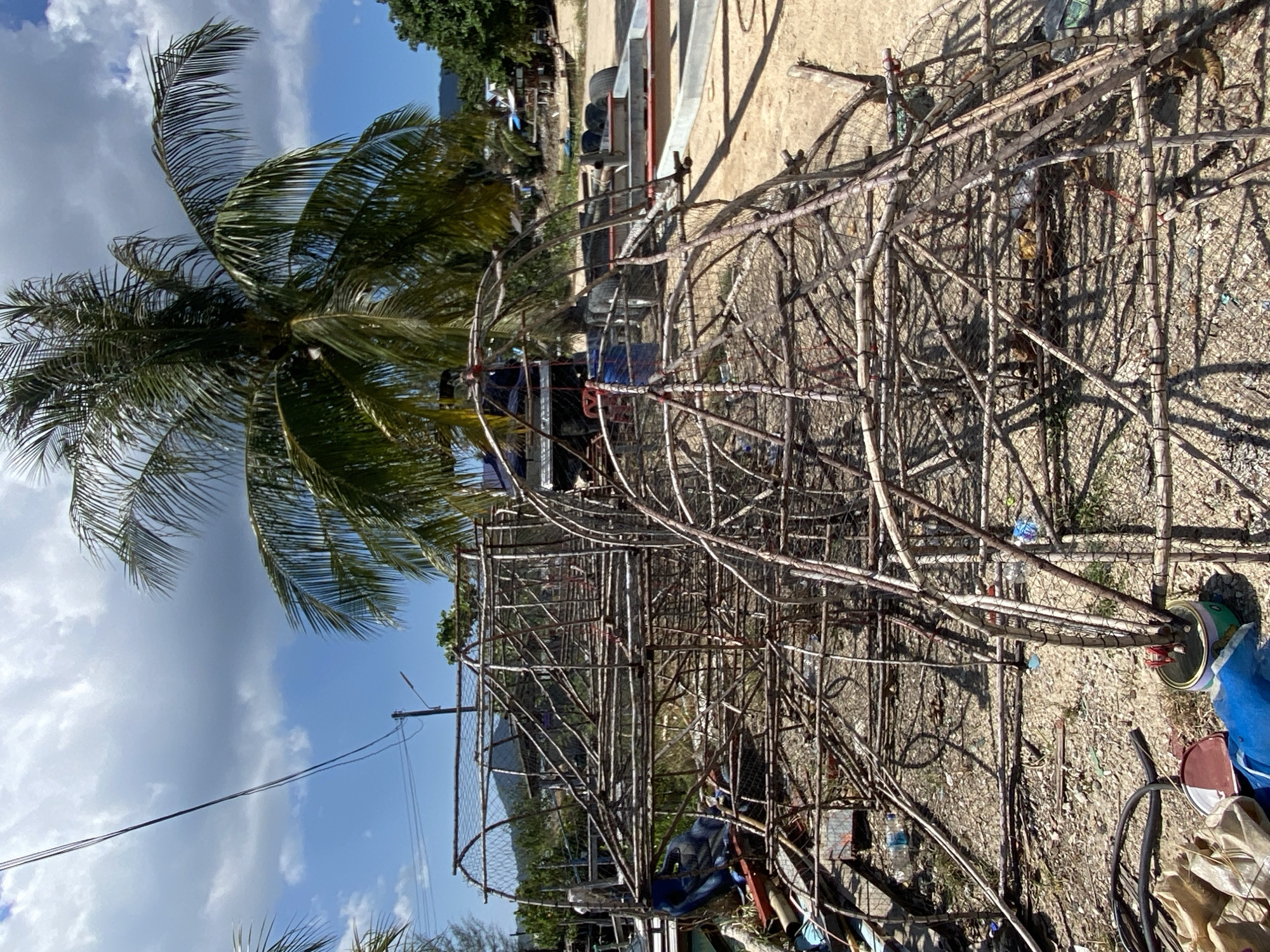 This media file captures an outdoor scene under bright midday sun, likely in Baan Wok Tum, Thailand. The primary subjects are numerous large, intricate structures crafted from wooden branches and netting, resembling traditional fish traps or cages. These structures occupy the foreground and midground, scattered across dry, gravelly ground that is littered with various debris, including plastic bottles and bags.
A tall palm tree with vibrant green fronds stands prominently on the left, reaching towards a clear blue sky dotted with fluffy white clouds. In the background, behind the woven structures, a vehicle, possibly a truck or part of a boat trailer, is partially visible. Further in the distance, more trees and the faint outline of mountains complete the setting.
The strong, distinct shadows cast by the structures indicate direct sunlight, consistent with late morning or early afternoon. No discernible human activity is taking place, suggesting the structures are either stored, under construction, or awaiting repair. There is no visible, legible text within the image. The overall setting is a tropical, possibly coastal, rural area.