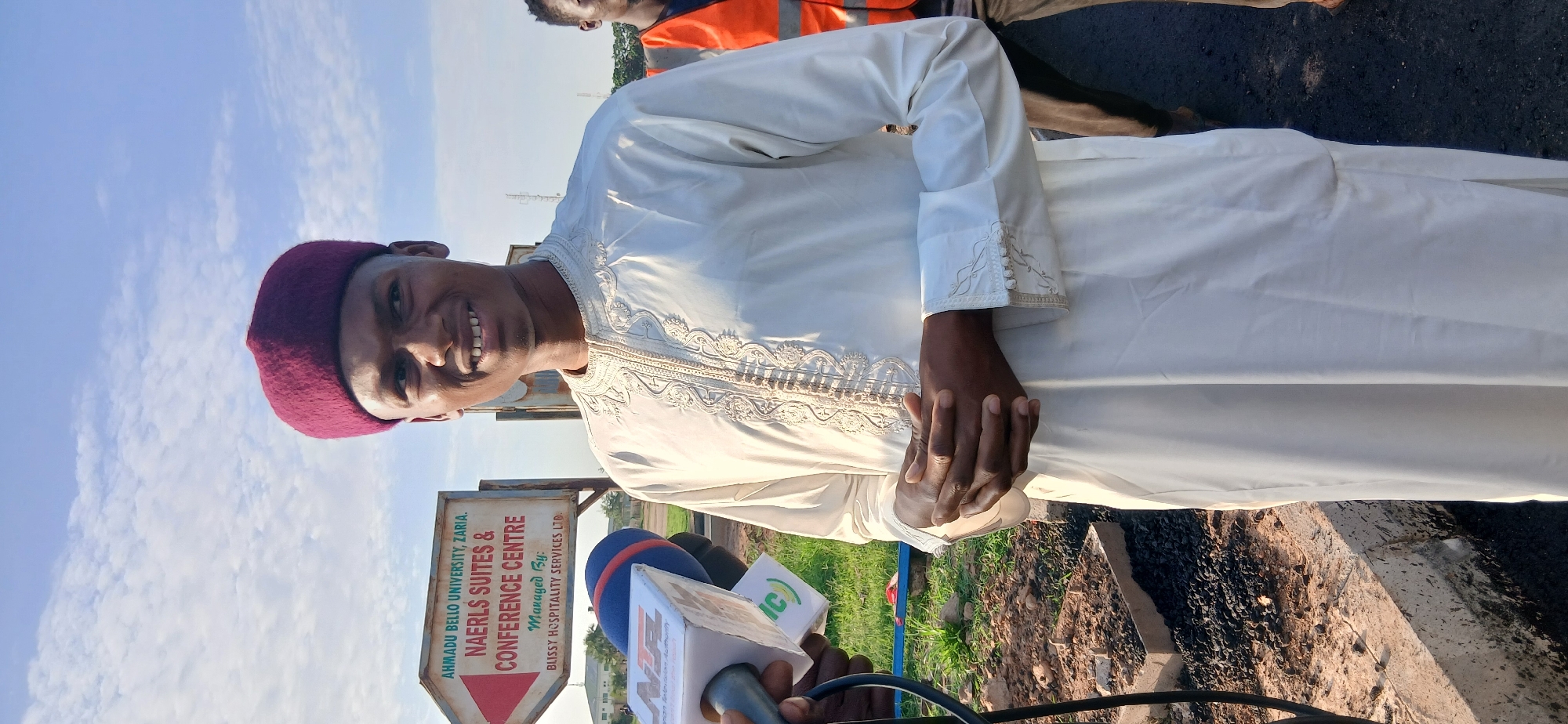 A young Black man, wearing a burgundy kufi and a white embroidered tunic, is being interviewed by a news crew in Jamaa A, Nigeria. He stands outdoors in front of a directional sign that reads "Ahmadu Bello University, Zaria. NAERLS Suites & Conference Centre. Managed By: Blissy Hospitality Services Ltd." The man is smiling and holding his hands clasped in front of him. A microphone with the Nigerian Television Authority (NTA) logo is visible, as is another microphone from a different news organization. The ground is covered with gravel and some small plants. In the background, another person wearing an orange safety vest is visible. The sky is partly cloudy.