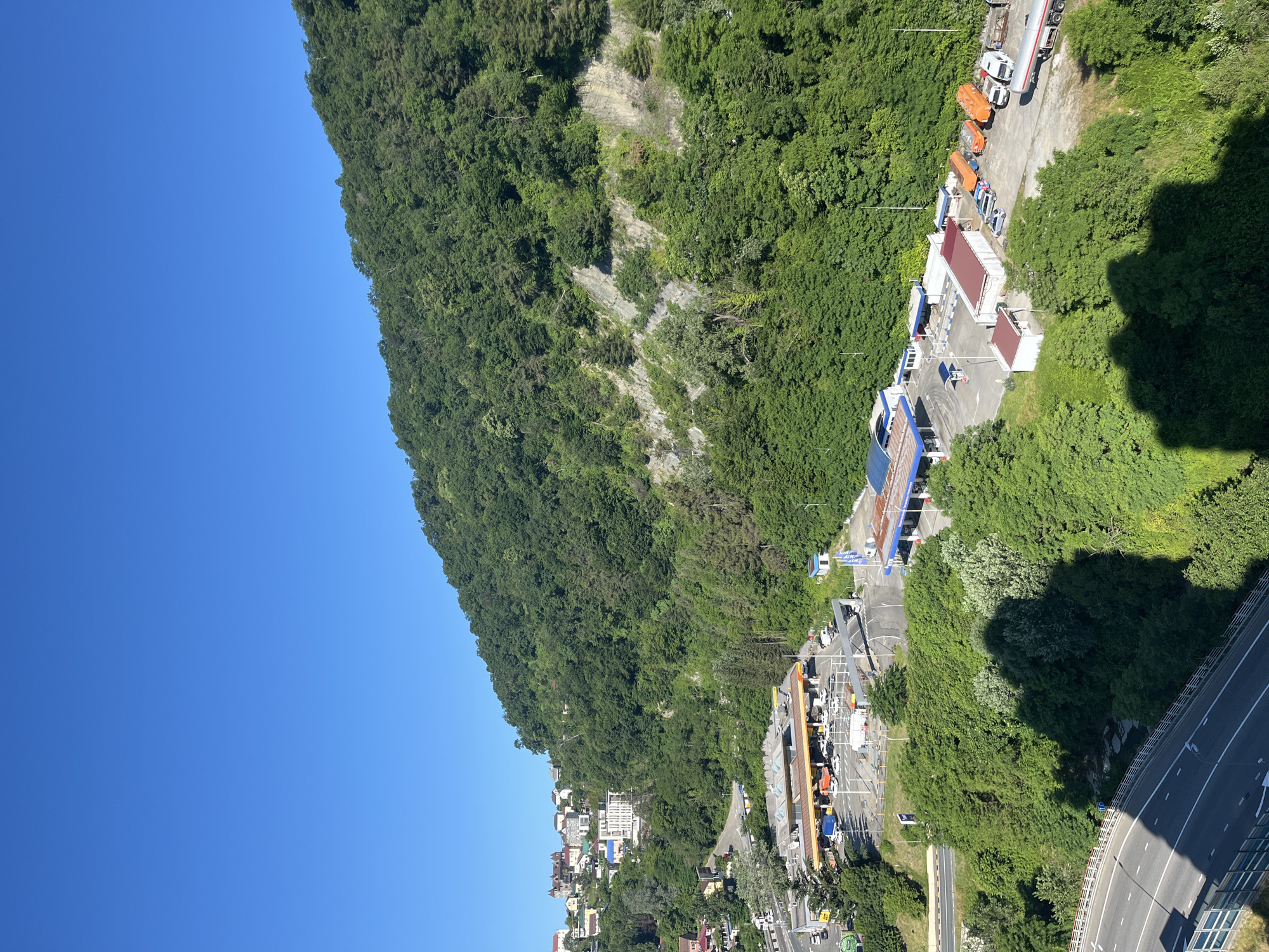 The image shows a view of the city of Sochi, Russia, with a prominent hill covered in dense green trees. In the foreground, a multi-lane road curves to the left, with a highway barrier along the right side. A large shadow of what appears to be a person is cast across the trees on the right side of the road.
Further into the scene, a cluster of buildings is visible on the side of the hill, appearing to be residential or commercial structures. Below these buildings and closer to the road, there is a gas station with multiple pumps and several vehicles. Adjacent to the gas station is another smaller complex of buildings, and further along the road, a toll plaza can be seen. The sky is a clear, vibrant blue, indicating a sunny day. The overall impression is a coastal city nestled against a lush, mountainous backdrop.