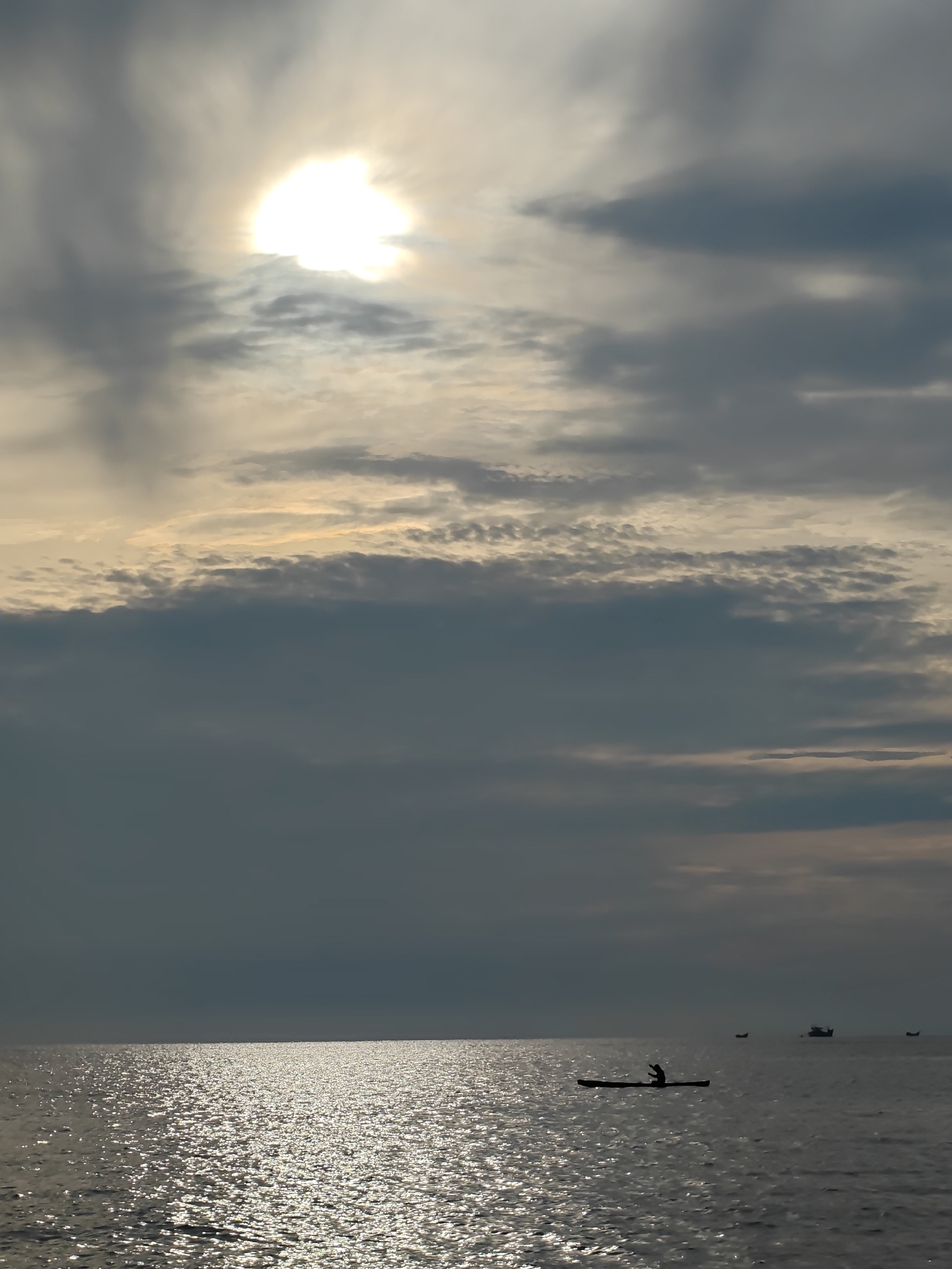 The image captures a serene seascape with a person kayaking in the foreground. The sun is partially obscured by clouds, casting a shimmering reflection on the water. In the background, several boats are visible, adding to the maritime atmosphere. The scene evokes a sense of tranquility and solitude, with the vast expanse of the ocean and the sky dominating the composition. The location is identified as Lhokseumawe, Indonesia, suggesting a tropical coastal setting.