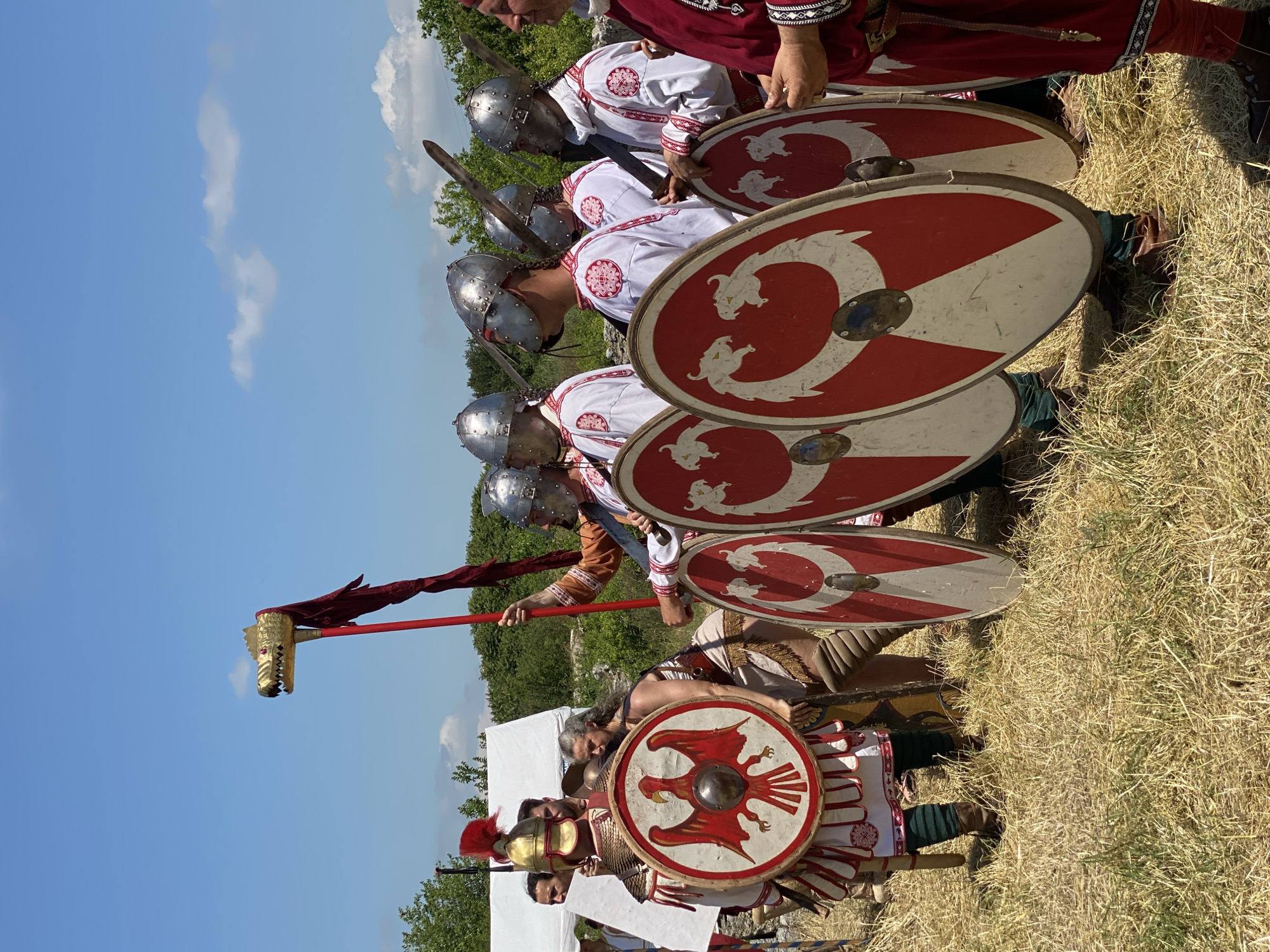 This image captures a historical reenactment event in Gigen, Bulgaria, under a clear blue sky with sparse white clouds. The ground is covered in dry, light brown grass.
In the foreground, a group of individuals dressed in historical attire stands. On the left, a person wearing a gold-colored segmented armor, a gold helmet with a red plume, and a white tunic with red trim, holds a large round shield featuring a red eagle on a white background. Behind them, partially obscured, is a woman with long gray hair, wearing a brown and cream tunic with fringe.
To the right, a line of several individuals is visible, dressed in white tunics with distinctive red circular patterns and wearing silver-colored helmets with face guards. Each of these individuals has a sword sheathed on their back/shoulder and holds a large round shield, which is red with an ornate white serpentine or dragon design. The person at the far right is partially cropped, wearing a red tunic with elaborate trim.
Between these two groups, a figure holds a tall red pole topped with a golden animal head, possibly a wolf or dragon (a draco standard), from which red fabric hangs. In the background, to the left, a white tent is visible, with dense green foliage and some scattered rocks beyond it. The scene is brightly lit by daylight.