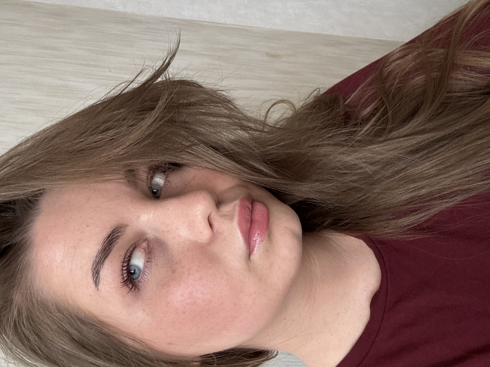 The image is a close-up portrait of a young woman with fair skin, light brown hair, and blue eyes. She is wearing a dark red top. Her eyebrows are well-defined, and she has light makeup on her lips. The background is a light, neutral color, possibly a wall. The image appears to be taken indoors. The image does not contain information about the location in the city of Chaikovsky, Russia.