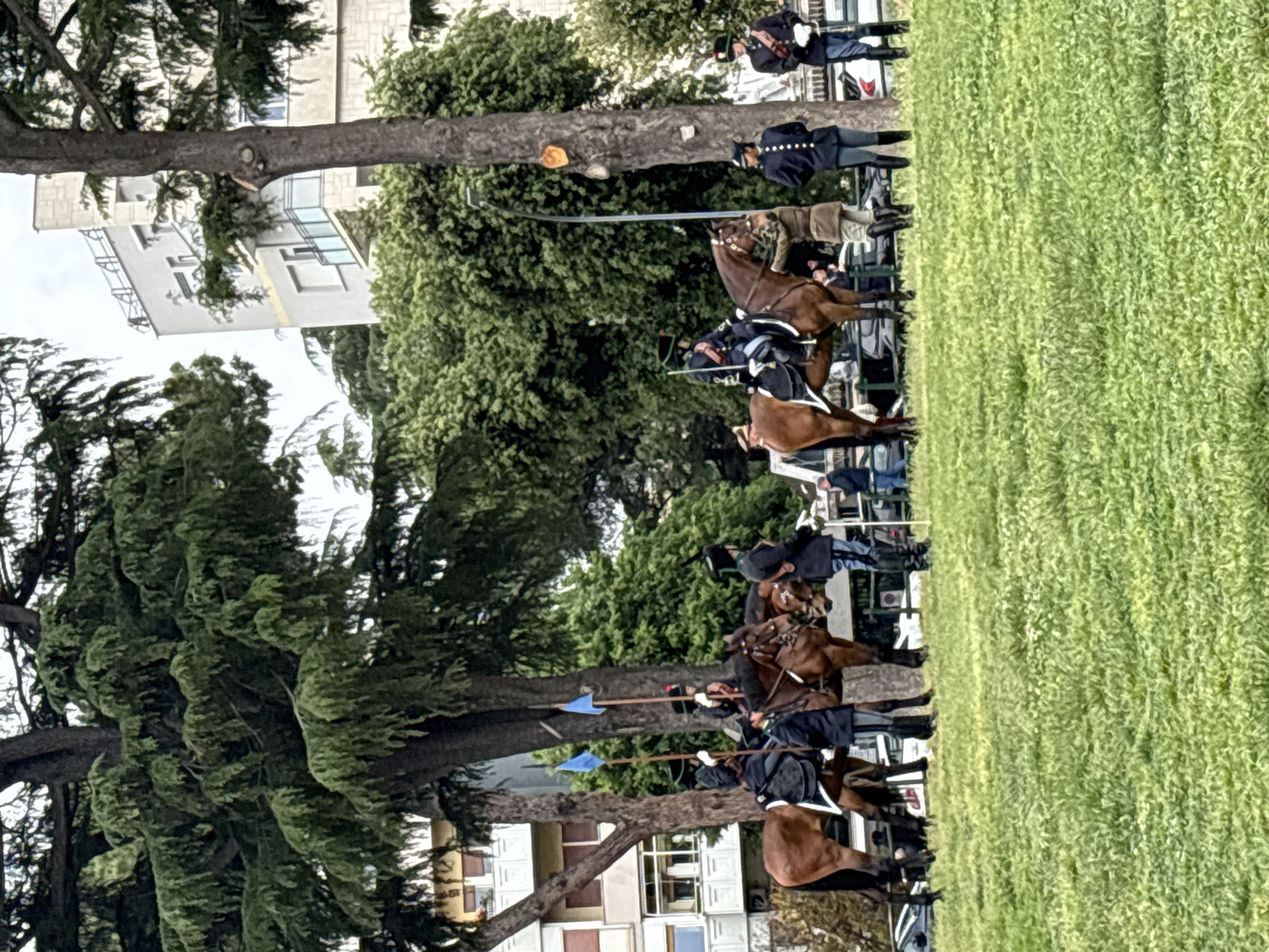 A parade of men on horseback, possibly re-enactors in historical military uniforms, proceeds across a green grassy area, likely a park or field within a city. The men wear dark-colored jackets with light breeches and tall black boots, holding long poles with blue flags or tips. The horses are primarily brown. Several spectators, dressed in modern clothing, stand along the sidelines. Trees and buildings are visible in the background, including a multi-story building with rectangular windows, suggesting an urban setting, specifically, a parade of the Roman Lanceri di Montebello on the Colle del Gianicolo (Janiculum Hill) in Rome, Italy, which is also visible in the background.