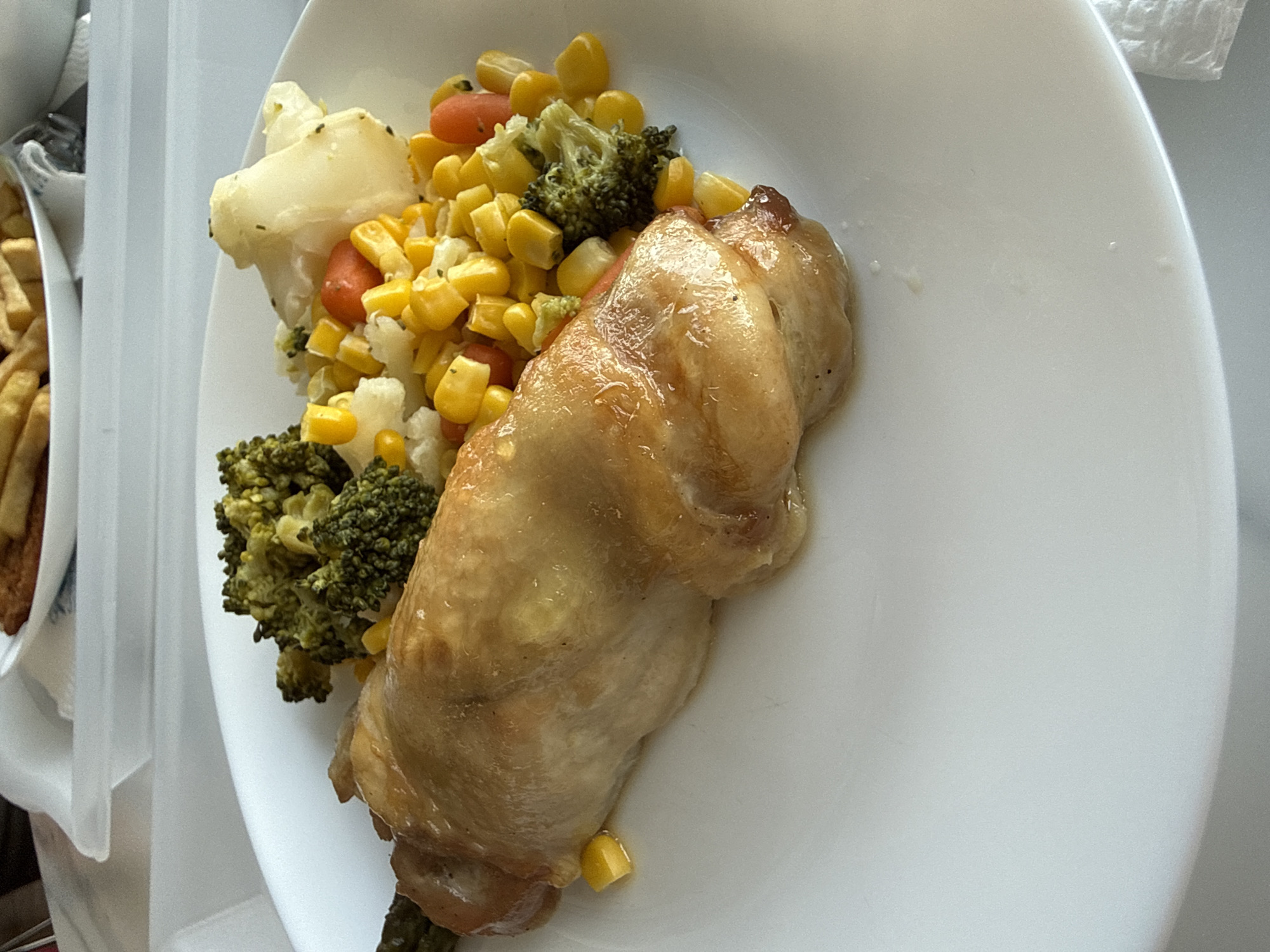 A white plate holds a portion of cooked chicken next to a mixture of steamed vegetables, including broccoli, cauliflower, corn kernels, and sliced carrots. The food appears to be on a table inside or near a tray holding a container of fries. There is no information indicating this photograph was taken in Sofia, Bulgaria.