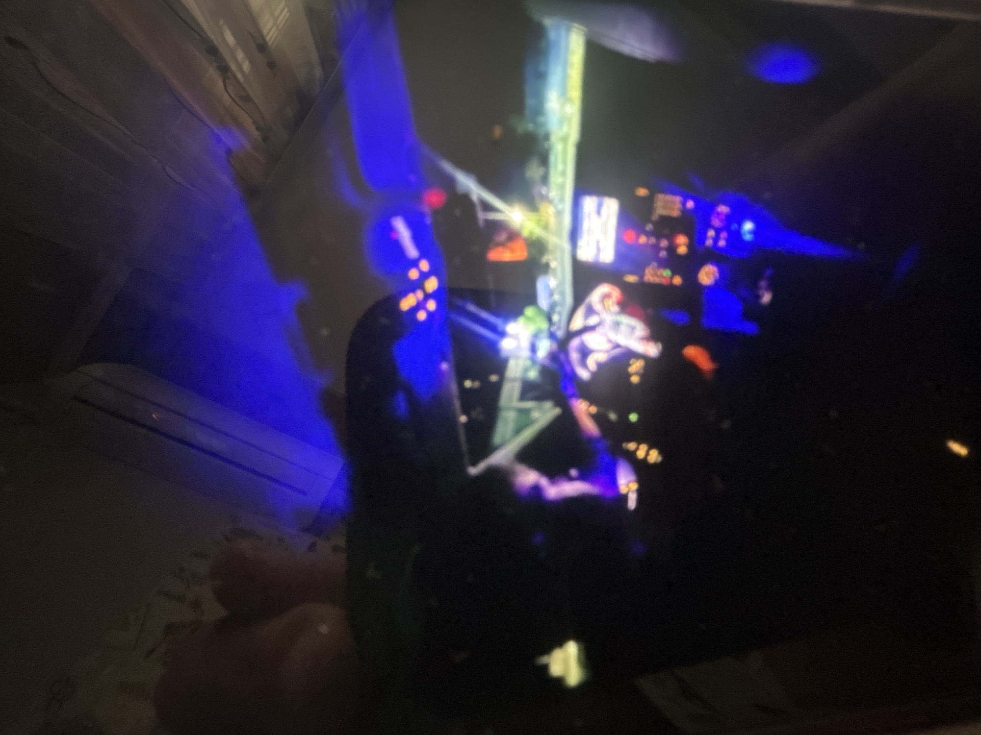 The image shows a photograph of a reflection of a video game being displayed on a screen. The room appears to be dimly lit with bluish lighting. The reflection is of a plane simulation video game with visible instruments. The location in the city is unknown.